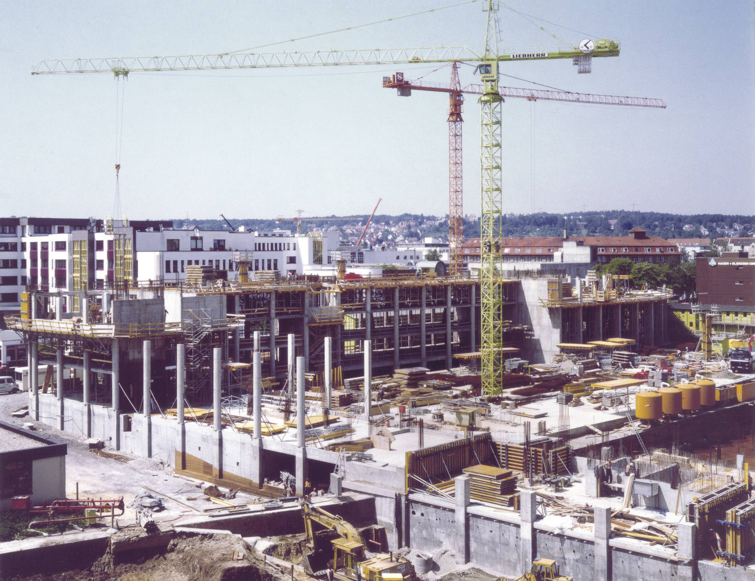 Un chantier de construction avec de grandes grues et un bâtiment multi-étages en cours de construction. Des ouvriers et des matériaux sont visibles.