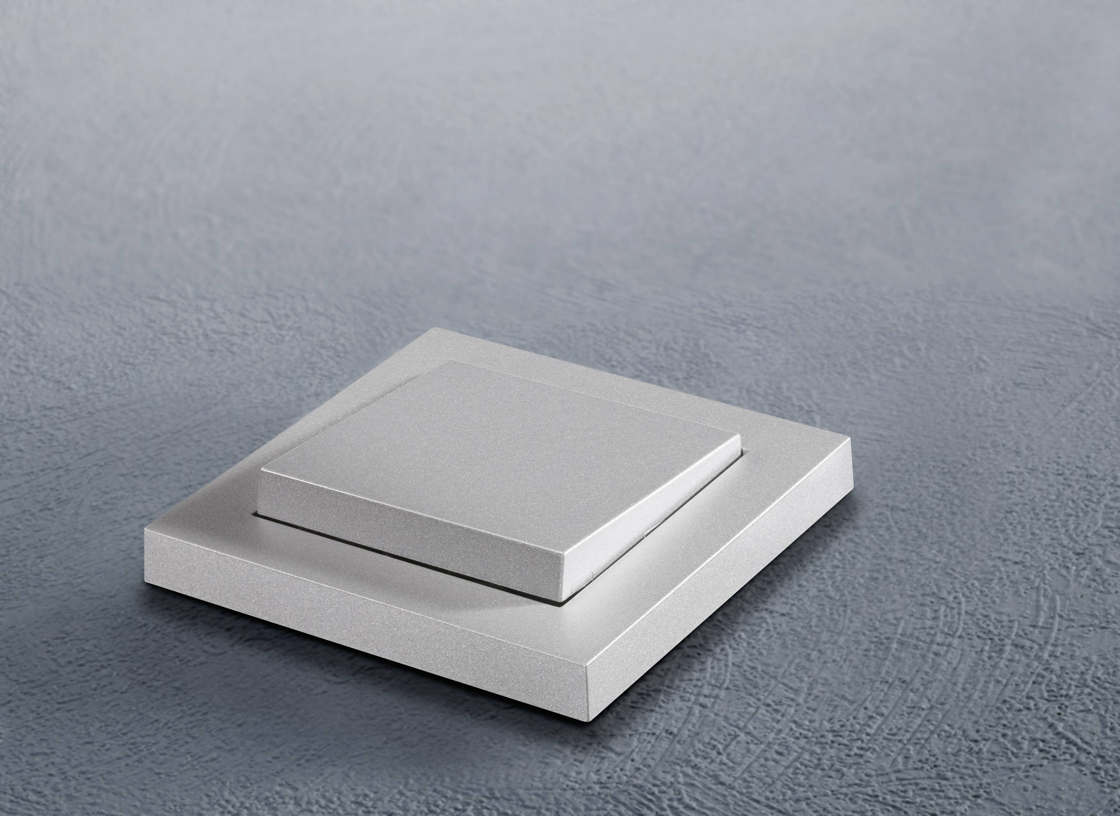 Un interrupteur argenté sur un mur gris texturé. L'interrupteur est carré et au design minimaliste.