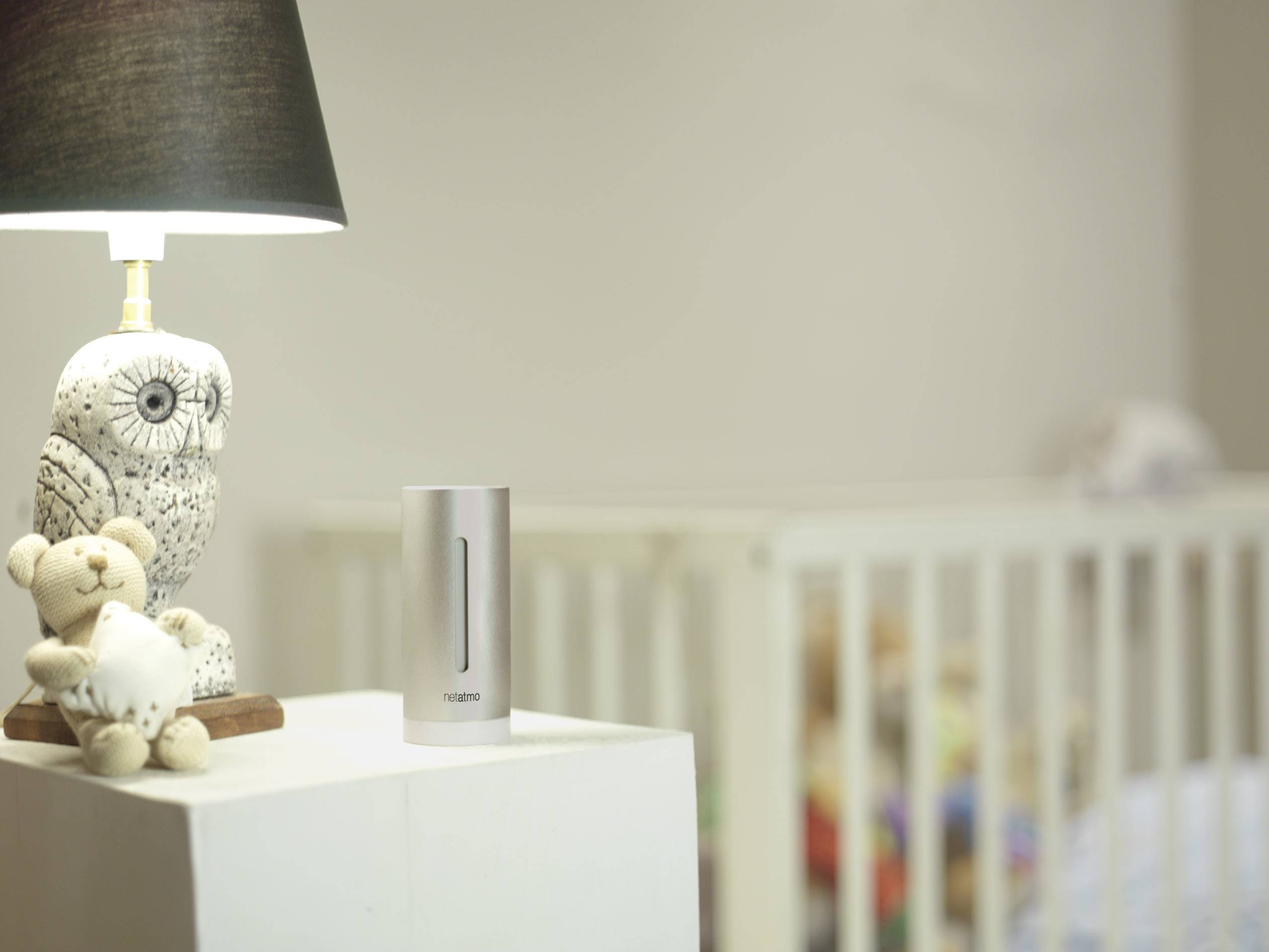 Une chambre d'enfant avec une table de chevet sur laquelle sont posés une lampe avec une figurine de hibou, un ours en peluche et un appareil numérique ; un lit de bébé se trouve en arrière-plan.