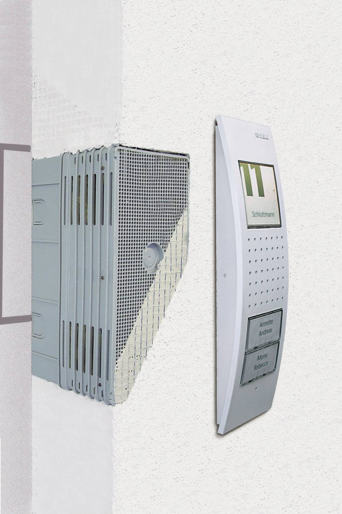 Un interphone extérieur blanc et moderne monté sur un mur clair. À sa droite, un dispositif visible avec des fentes de ventilation.