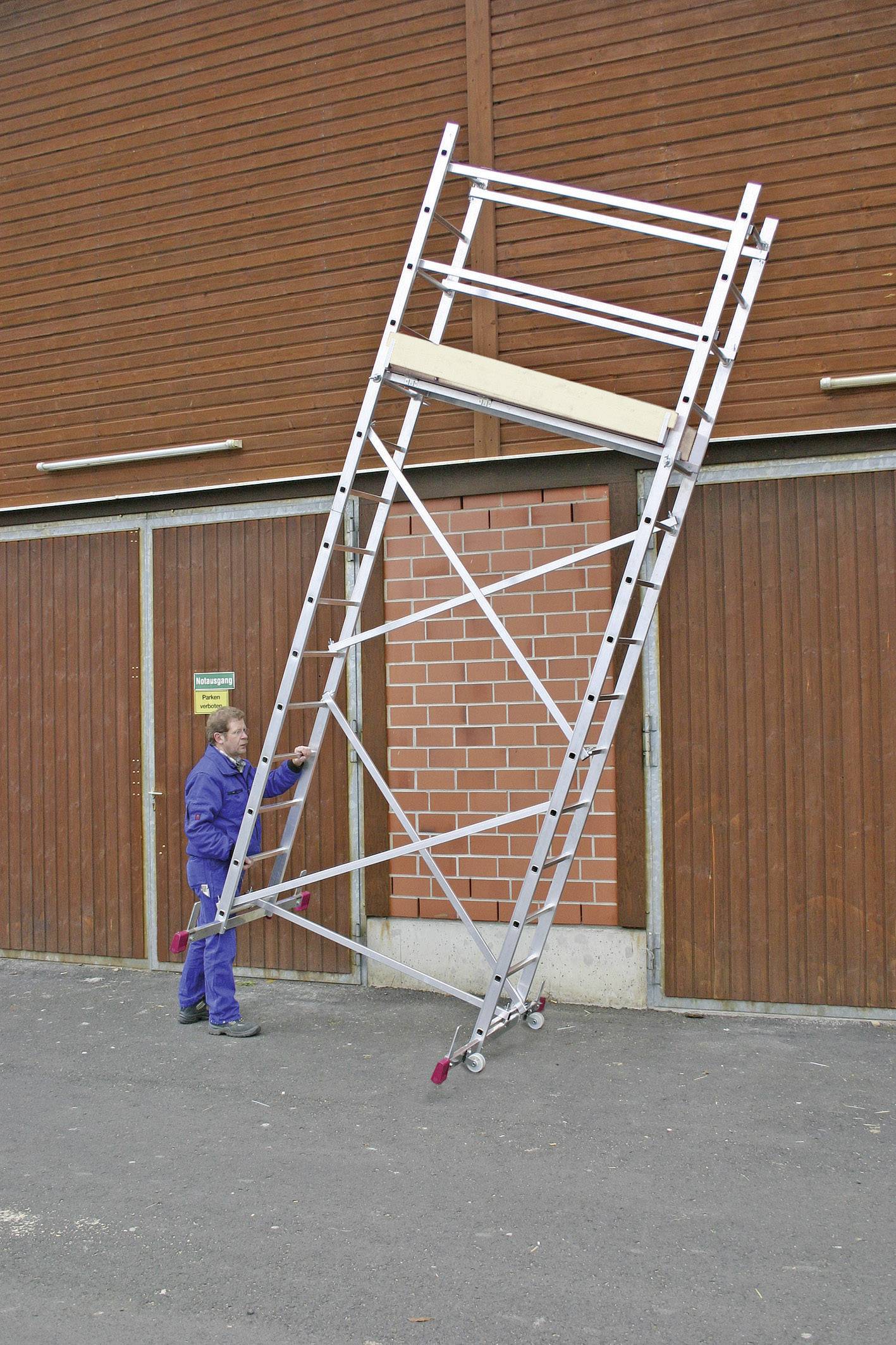 Une personne en tenue de travail tient un grand échafaudage incliné sur une cour pavée devant un mur en bois rouge.