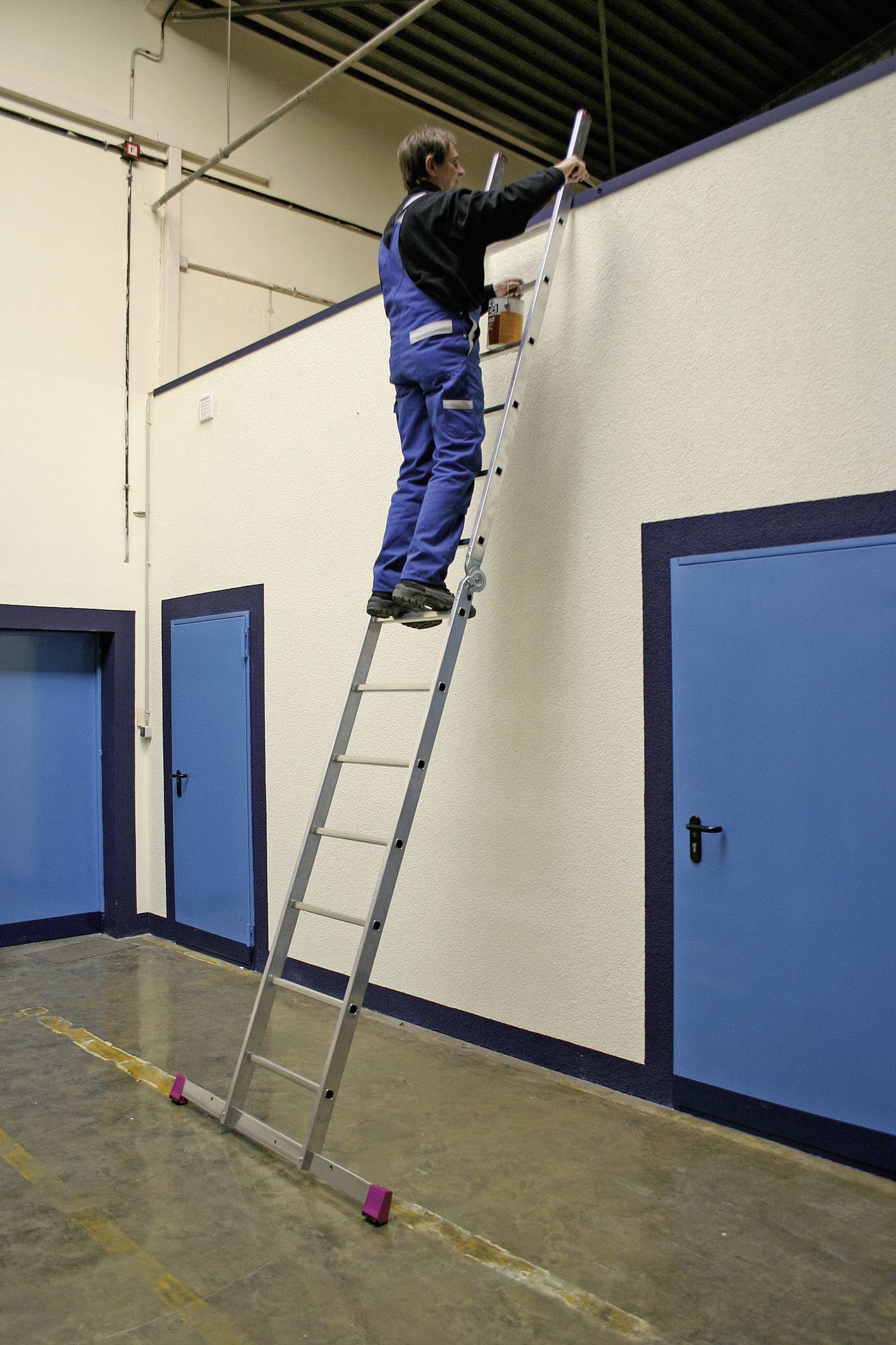 Une personne se trouve sur une échelle et peint le mur supérieur d'une pièce. Les murs ont des portes bleues.