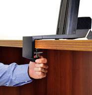 Une personne fixe le pied d'un support de bureau à l'aide d'un bouton de serrage à main sur un bureau en bois. Le bureau est brun foncé.