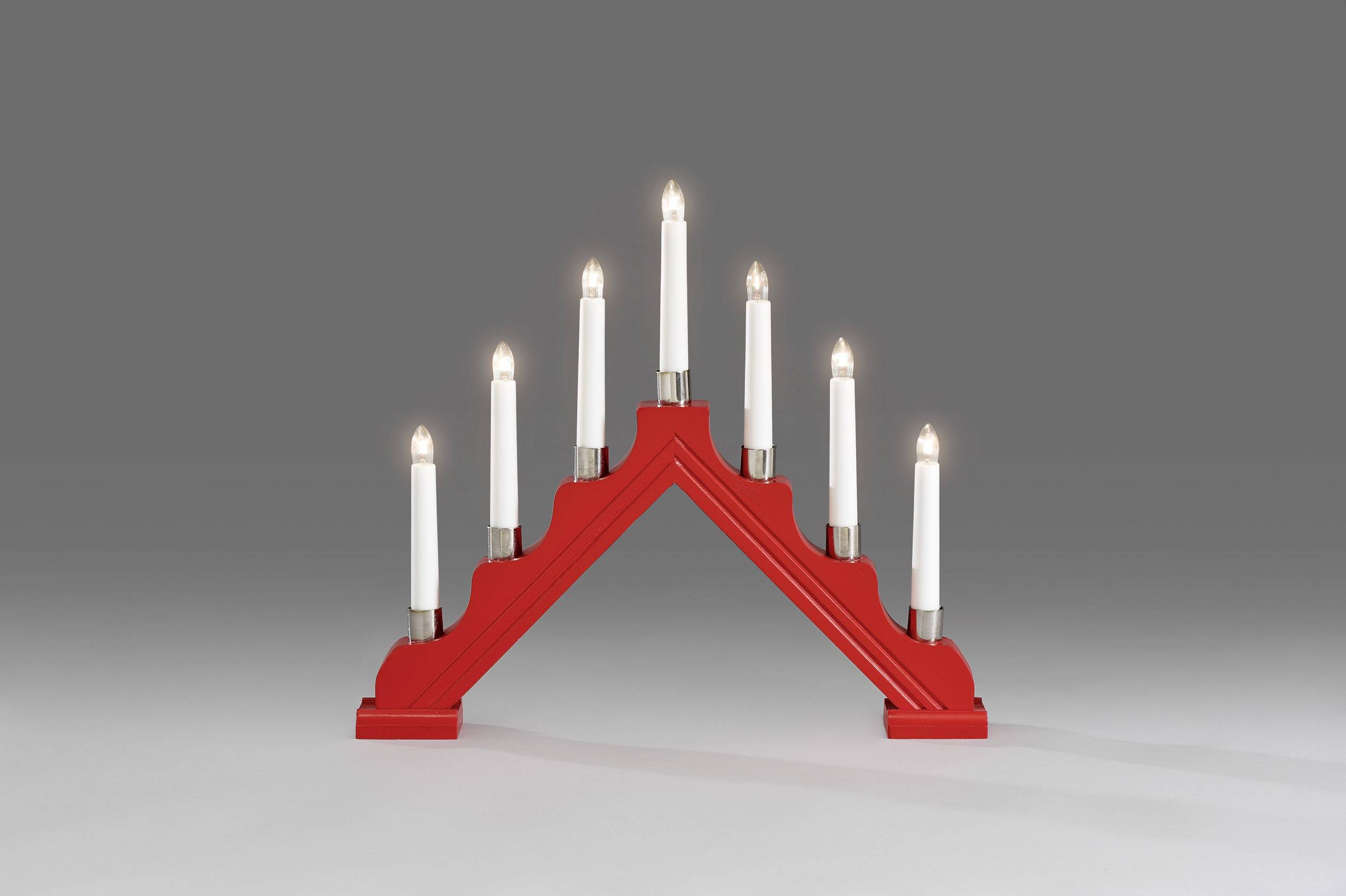 Un support de bougie rouge triangulaire avec sept bougies blanches allumées, posé sur une surface grise devant un fond gris.