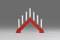 Un support de bougie rouge triangulaire avec sept bougies blanches allumées, posé sur une surface grise devant un fond gris.