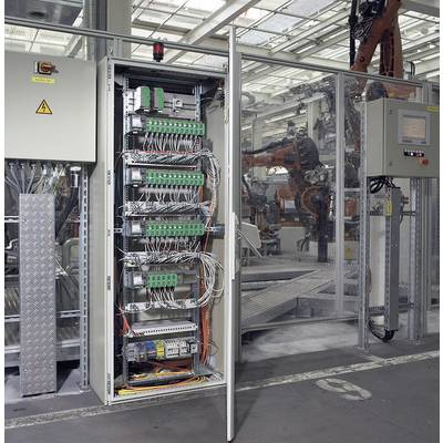Un armoire électrique ouverte montre une multitude de câbles et de composants électriques. En arrière-plan, on aperçoit une installation de fabrication.