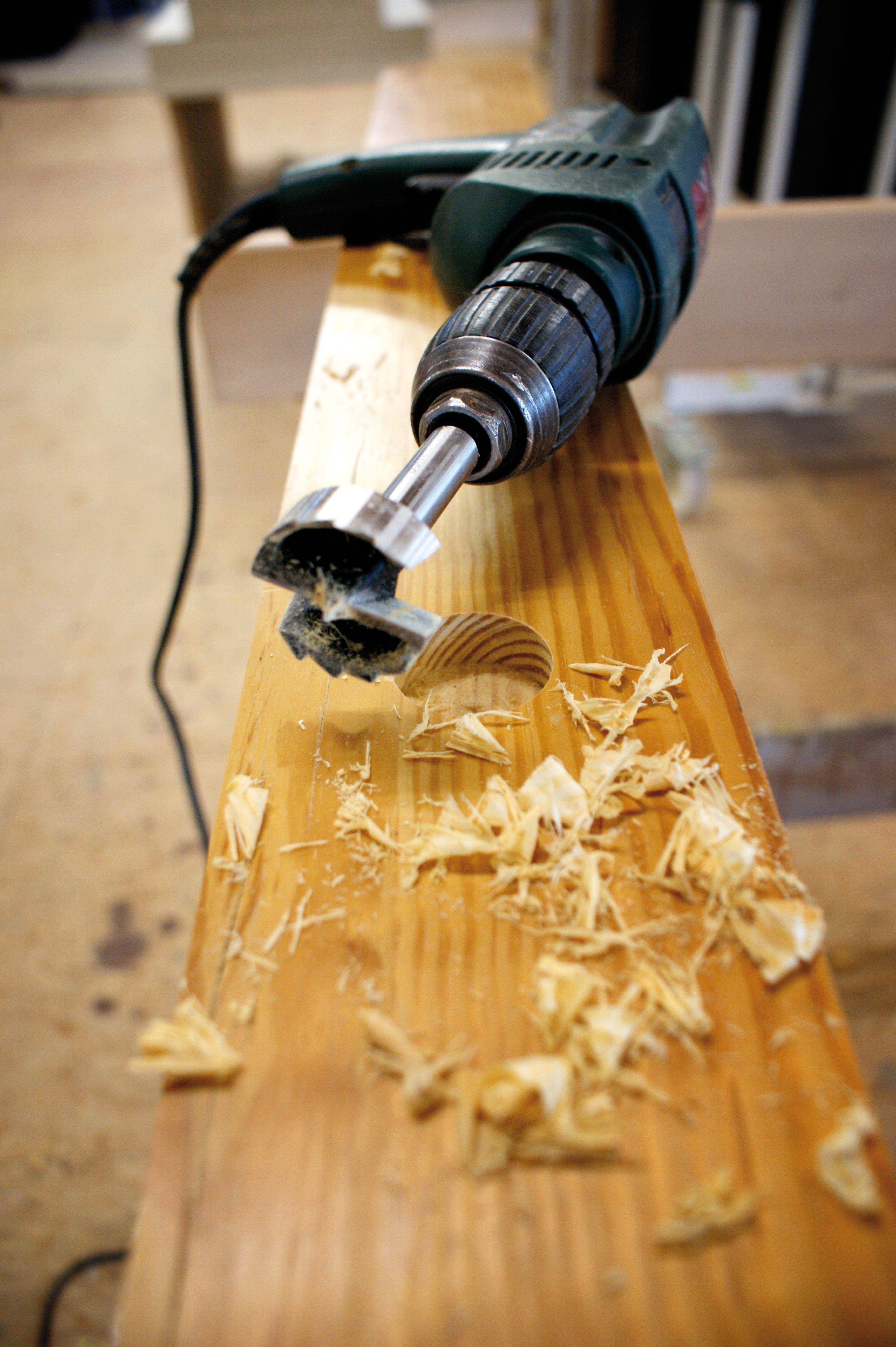 Une perceuse avec un mèche à façonner est posée sur une table en bois. Des copeaux de bois sont dispersés autour de la mèche.