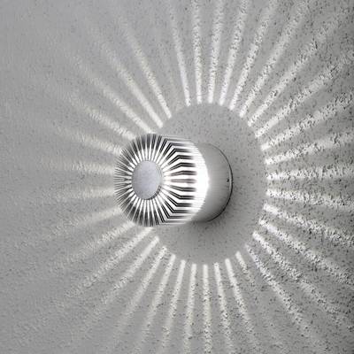 Un applique cylindrique projette un motif lumineux rayonnant sur le mur, évoquant des rayons de soleil.