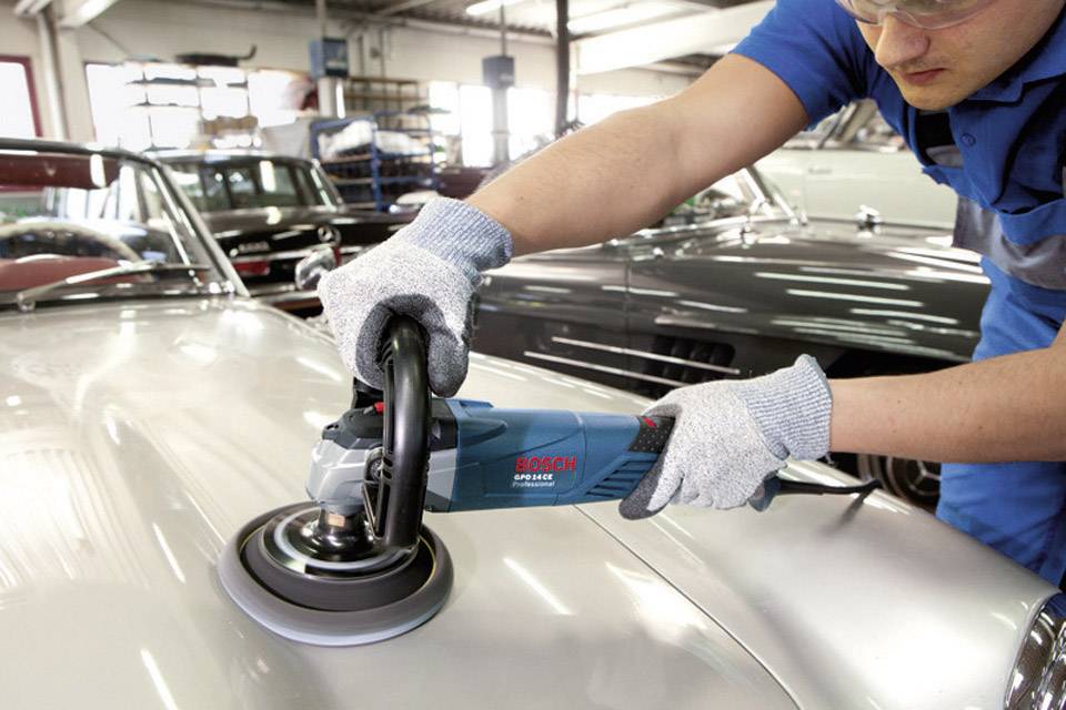 Une personne polit le capot d'une voiture argentée à l'aide d'une polisseuse électrique dans un atelier.