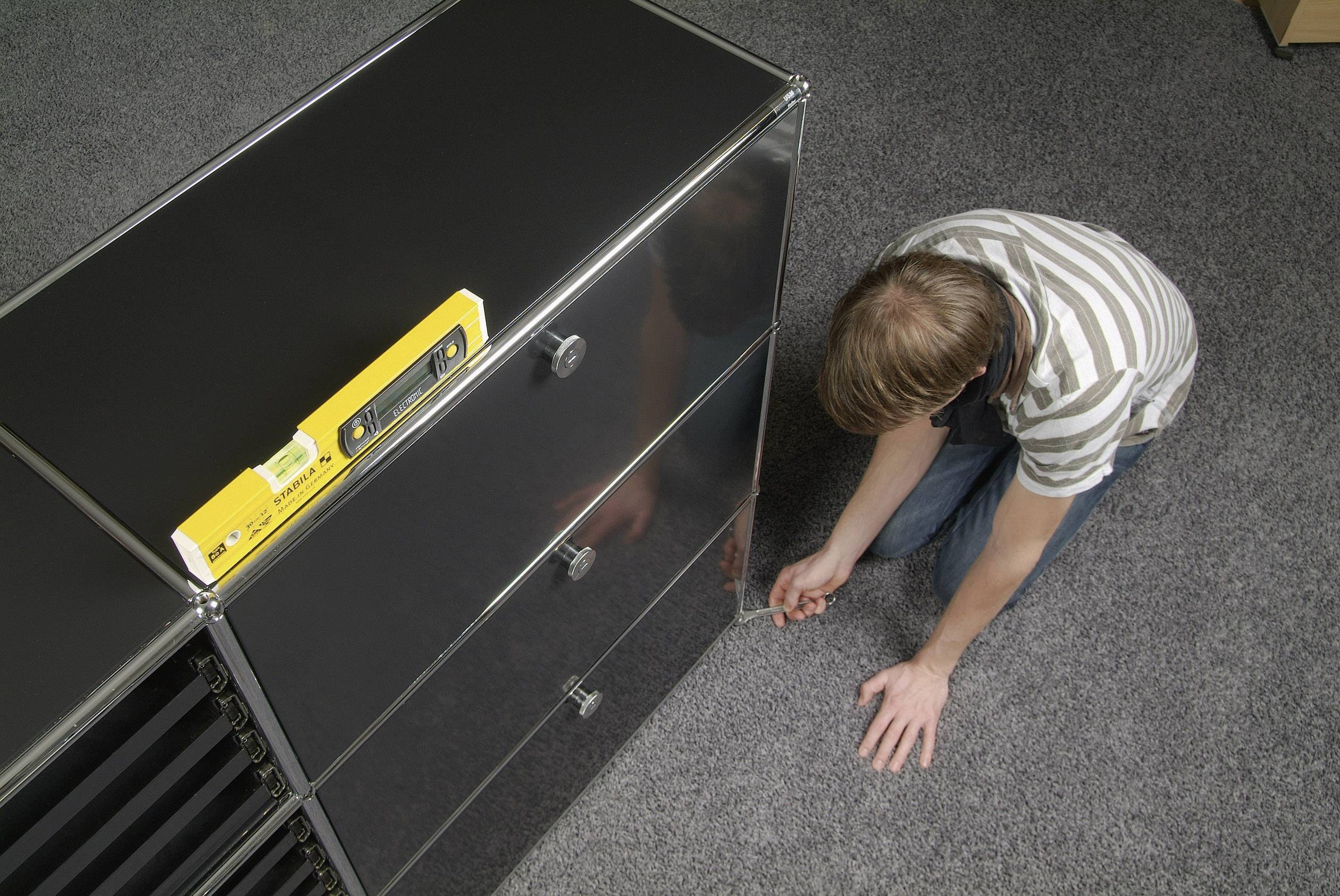 Une personne portant un t-shirt rayé est agenouillée au sol et travaille sur un meuble noir. Un niveau à bulle est posé dessus.