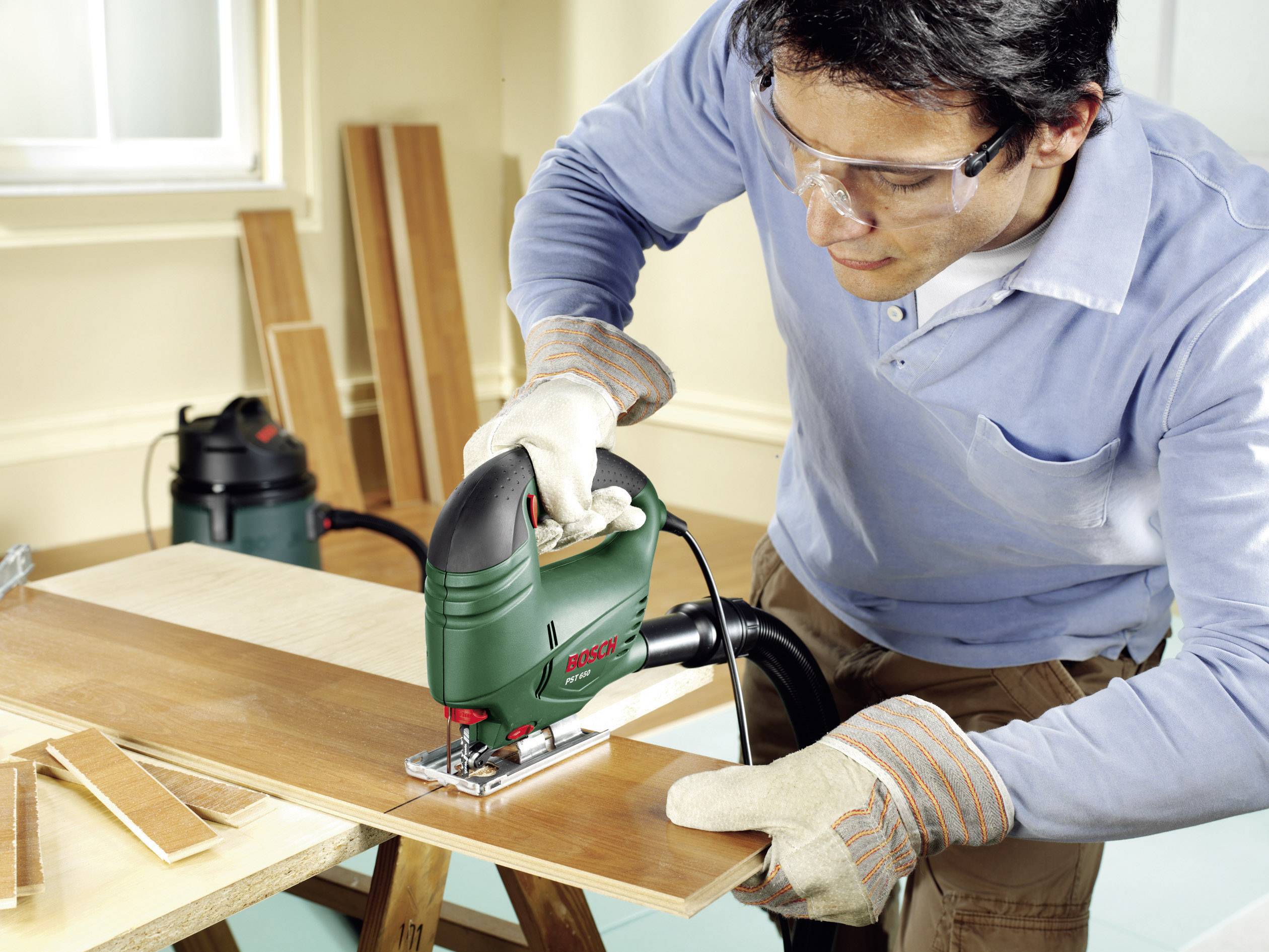 Un homme portant des lunettes de protection et des gants utilise une scie sauteuse électrique pour couper une planche en bois. Des planches sont visibles en arrière-plan.