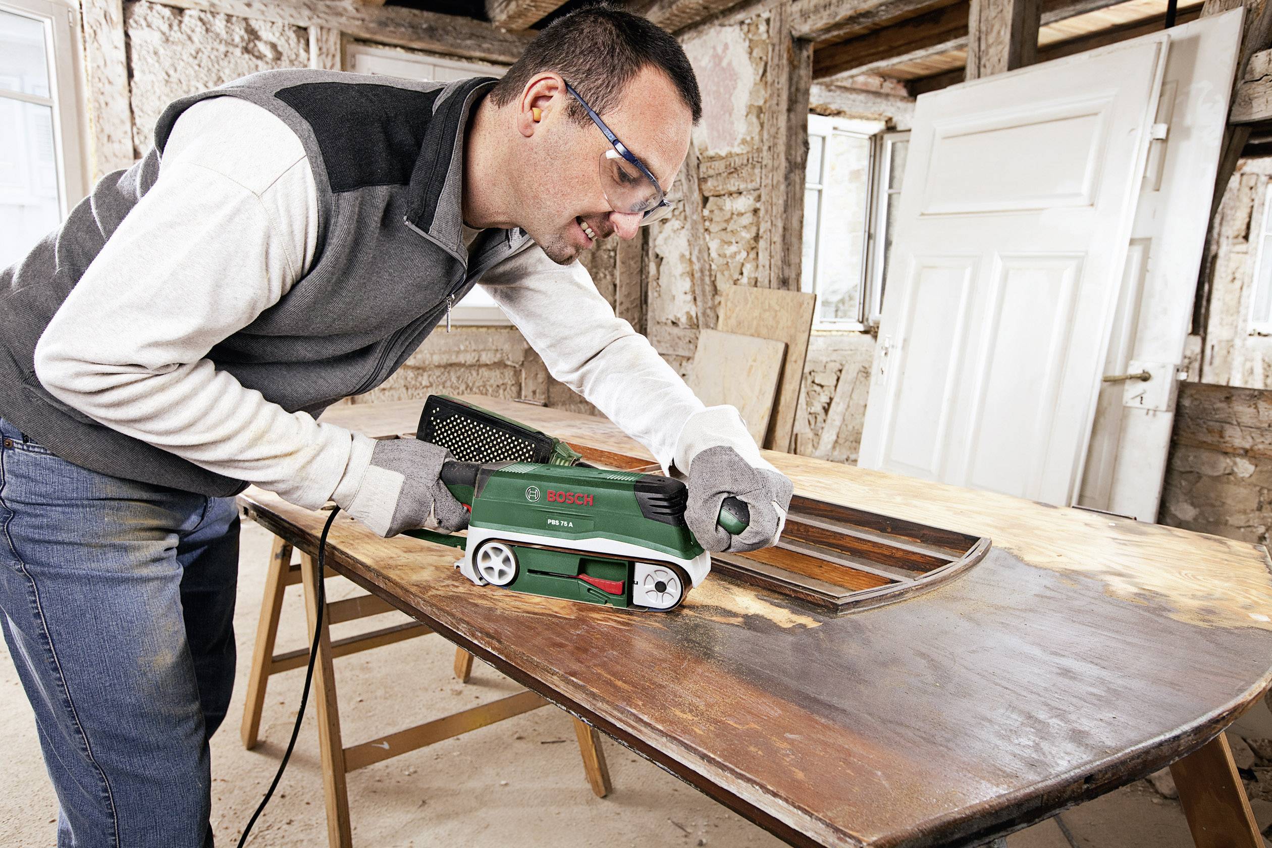 Une personne ponce un grand plateau de table en bois avec une ponceuse dans un atelier. Elle porte des lunettes de protection et des gants.