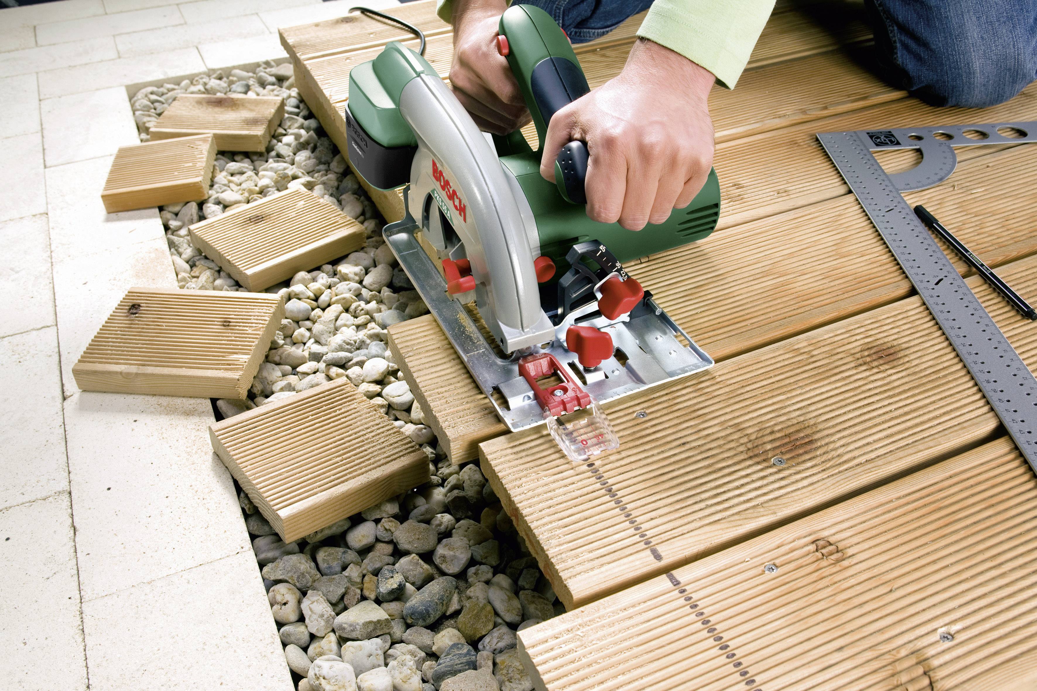 Une personne scie du bois avec une scie circulaire sur une terrasse. Des copeaux de bois et du gravier en arrière-plan soulignent le contexte de bricolage.