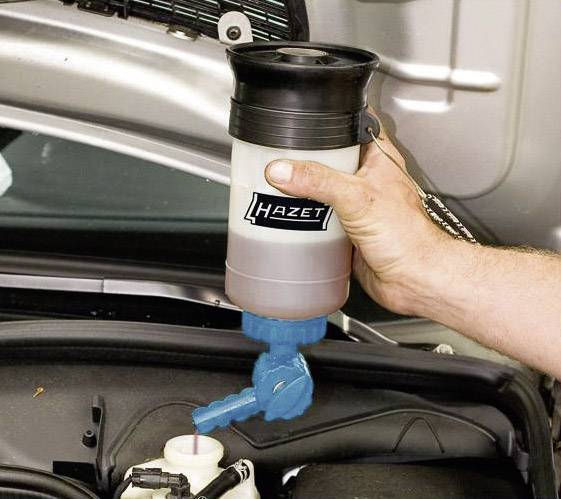 Une main tient un distributeur de liquide au-dessus du moteur d'une voiture. Le distributeur a une valve bleue et le logo 'Hazet'.
