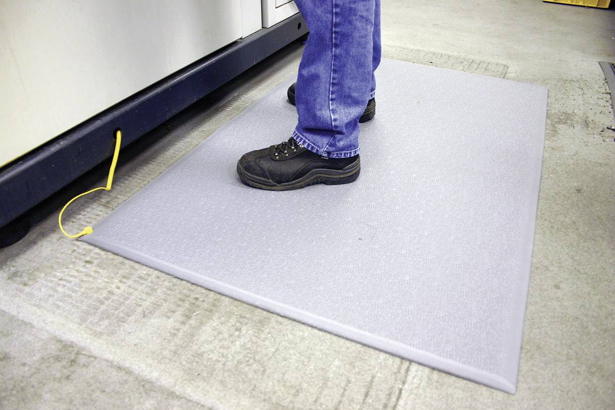 Une personne se tient sur un tapis anti-fatigue gris devant une machine. Le tapis a un câble jaune pour la mise à la terre.