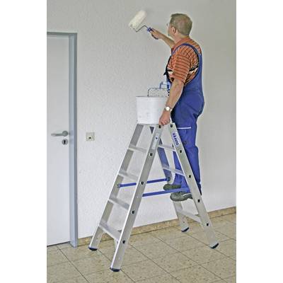 Un homme en salopette bleue est debout sur une échelle et peint le mur en blanc avec un rouleau à peinture.