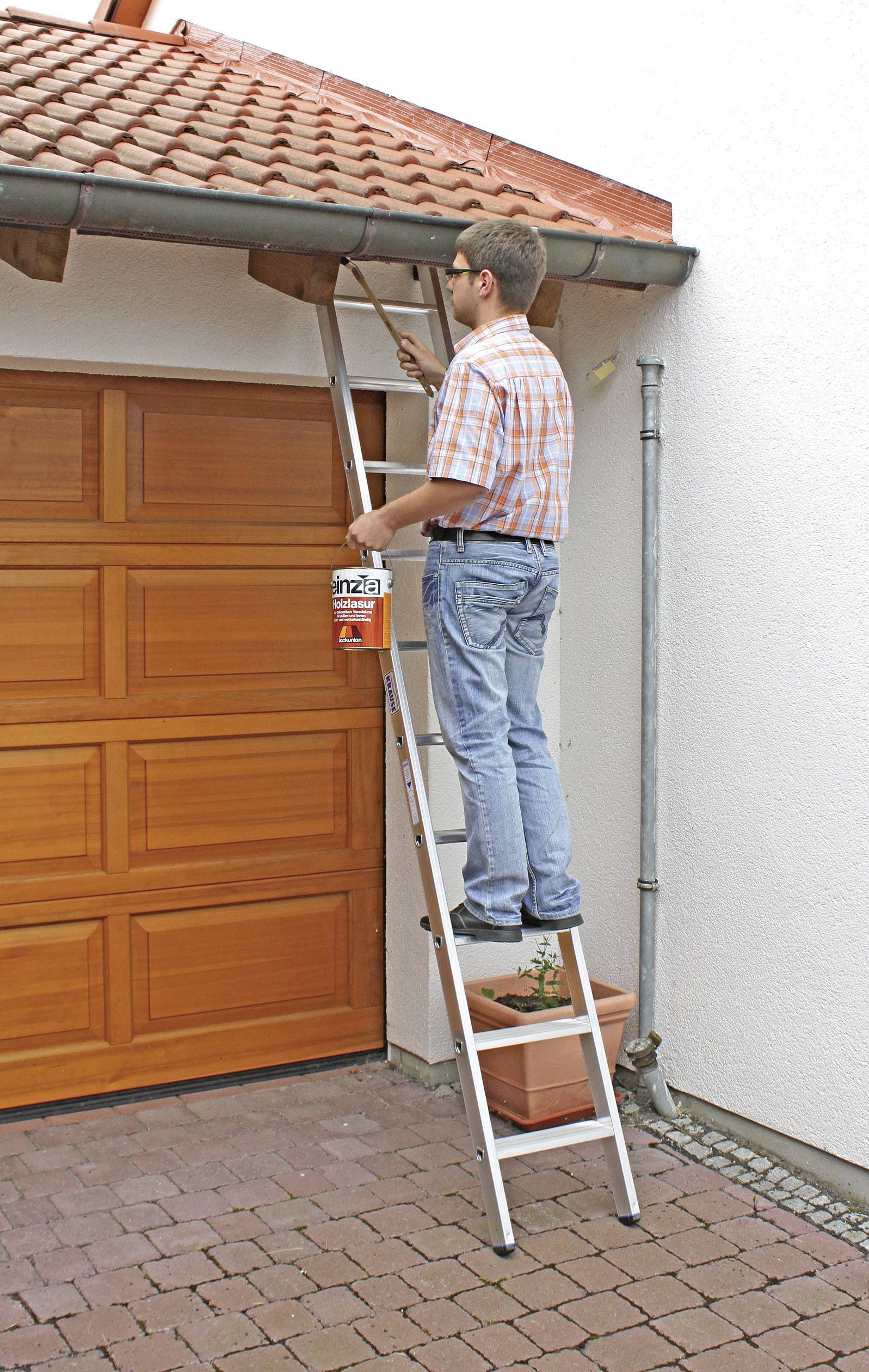 Une personne se trouve sur une échelle et peint la gouttière d'une maison. Un portail en bois est visible à gauche.