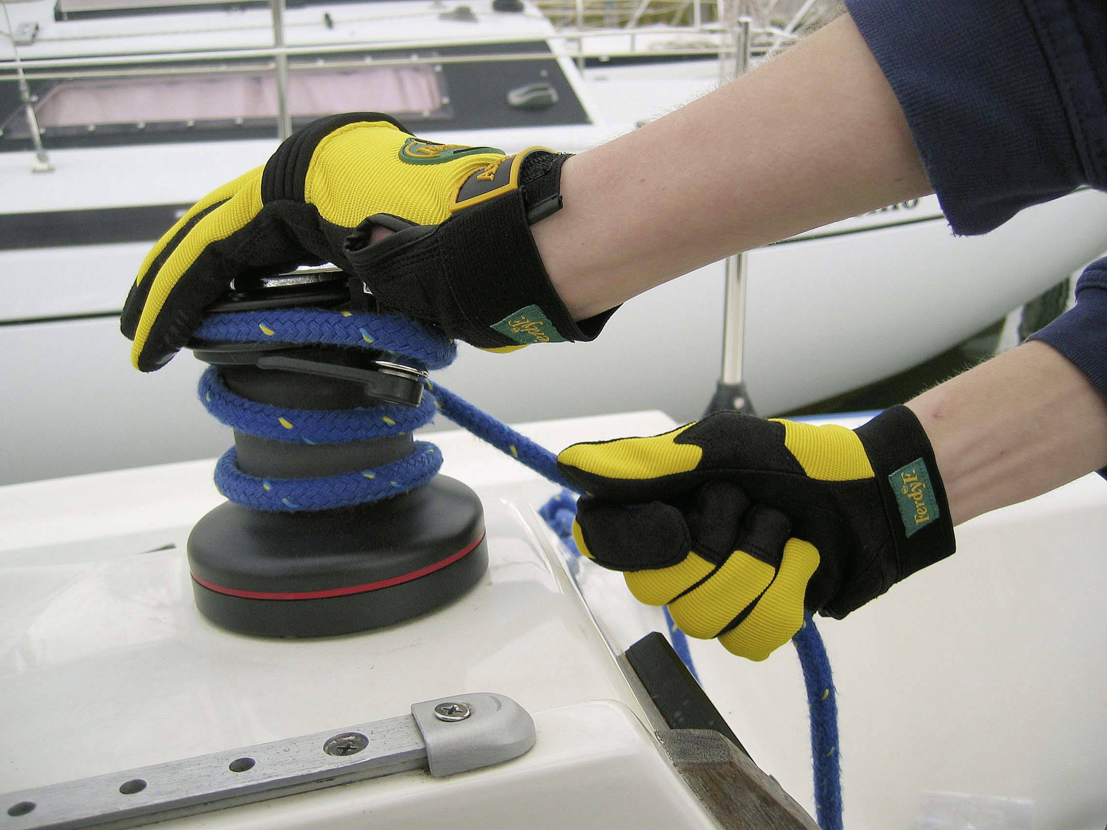 Deux mains gantées de jaune manipulent un cordage sur un winch d'un voilier. On distingue des parties du bateau en arrière-plan.
