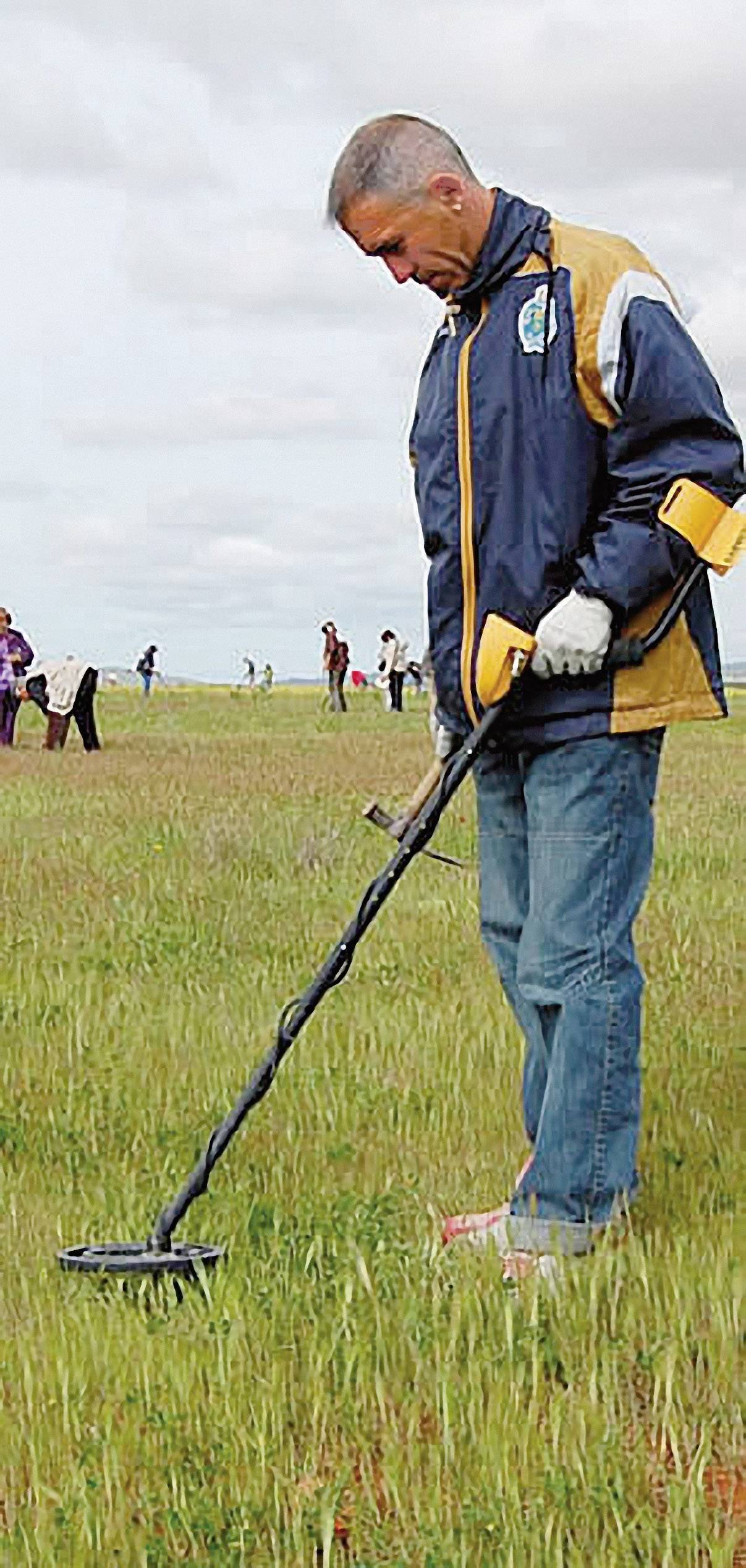 Un homme avec un détecteur de métaux cherche des objets métalliques dans un champ. D'autres personnes sont floues en arrière-plan.