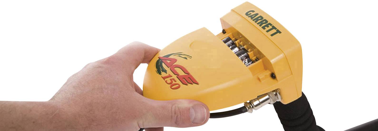 Une main tient un appareil jaune portant l'inscription 'ACE 150', utilisé pour la détection de métaux. 'GARRETT' est inscrit en haut. Un câble est connecté.