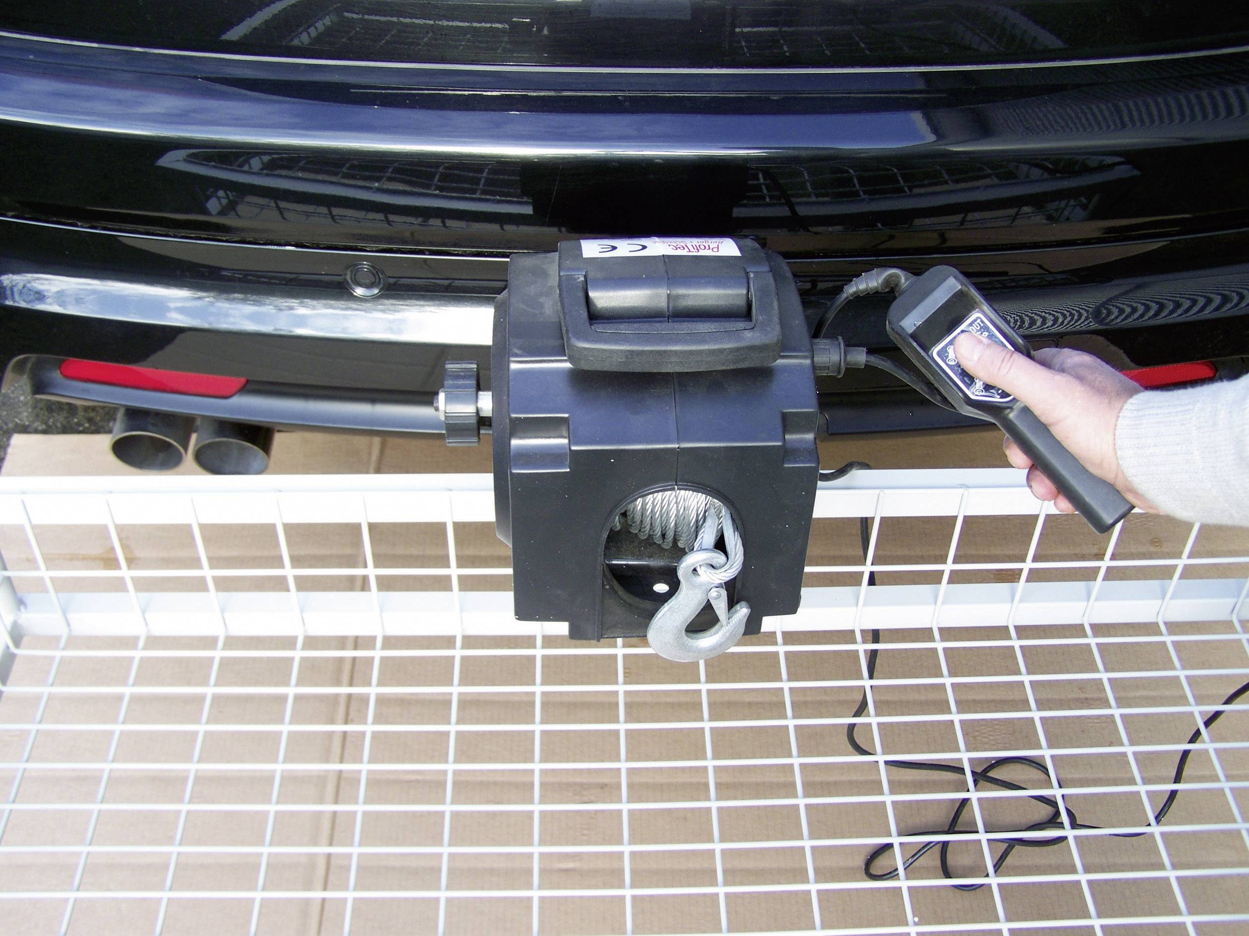 Un câble de traction est fixé à l'arrière d'une voiture. Une main tient une télécommande pour contrôler le câble de traction suspendu à un châssis grillagé.