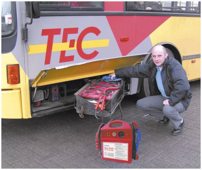 Un homme pointe du doigt le couvercle du compartiment à batterie ouvert d'un bus urbain. La batterie est partiellement extraite. Une unité de charge rouge est placée devant.