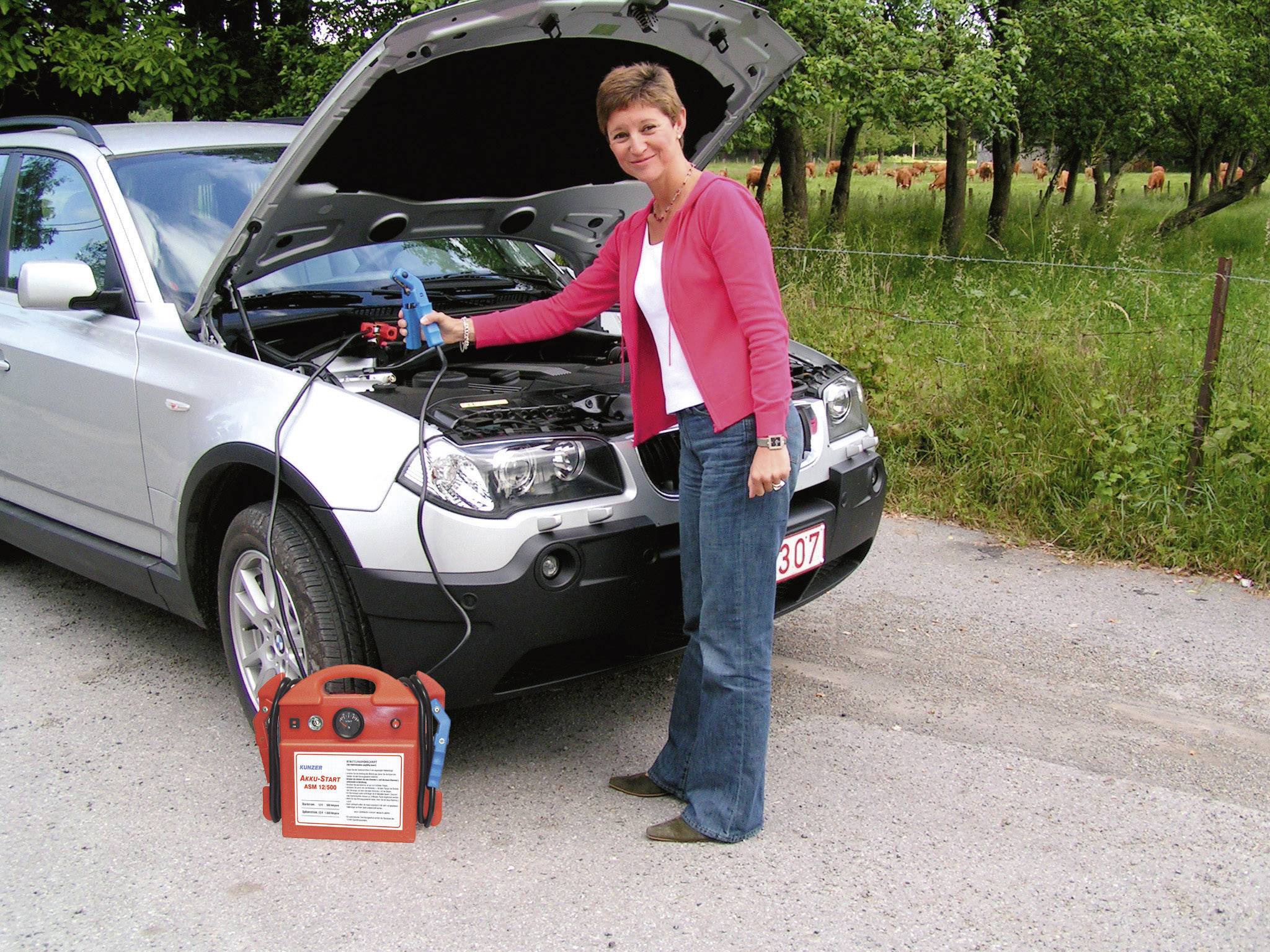 Une femme se tient devant une voiture, capot ouvert, et tient un câble de démarrage connecté à un chargeur de batterie.