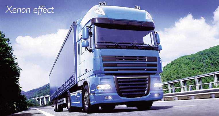 Un camion bleu roule pendant la journée sur une route avec un pont. Des arbres et le ciel sont visibles en arrière-plan. Texte : 'Effet xénon'.
