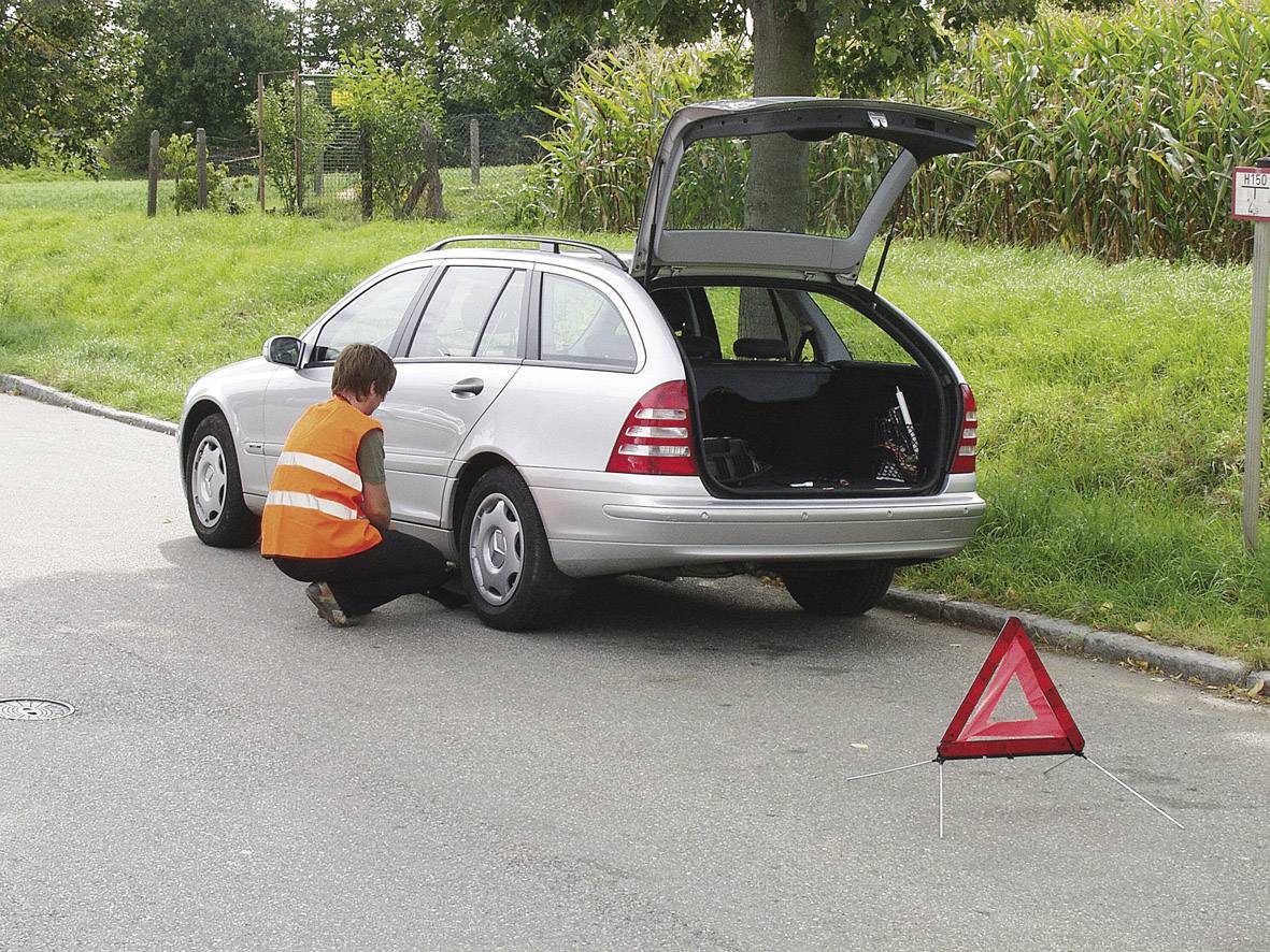 Une personne portant un gilet de sécurité change un pneu sur une voiture argentée au bord de la route. Un triangle de signalisation est installé.