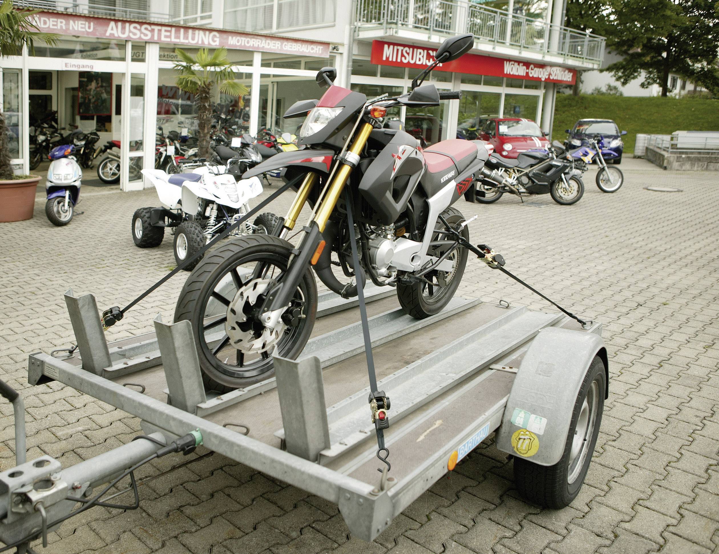 Une moto rouge est transportée sur une remorque devant la vitrine d'un concessionnaire de motos. D'autres motos sont visibles en arrière-plan.