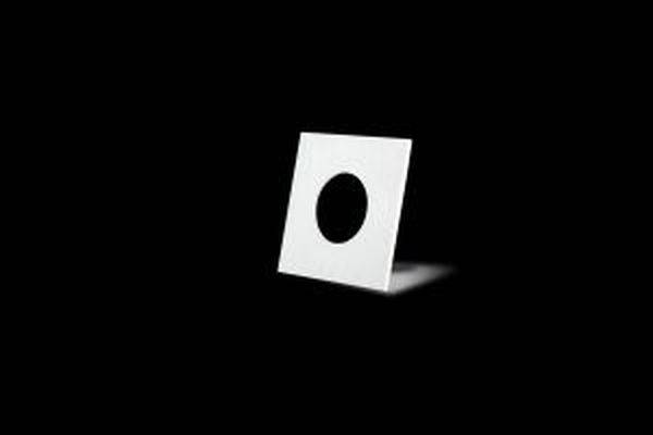 Une feuille blanche carrée avec un trou rond en son centre est légèrement inclinée sur une surface sombre devant un fond noir.
