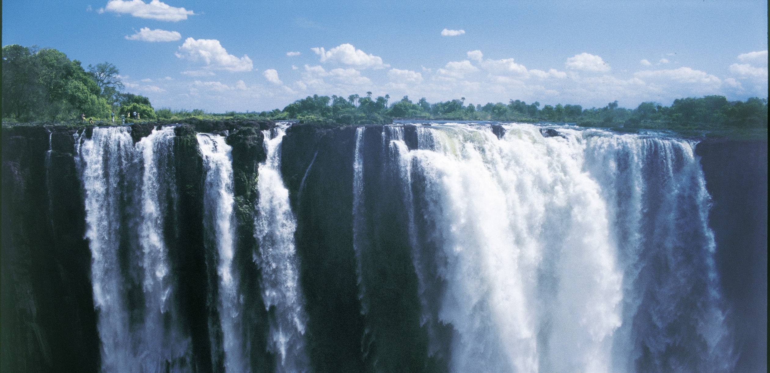 Des cascades au débit puissant se déversent dans un canyon, entourées de végétation verdoyante et d'un ciel bleu parsemé de nuages.