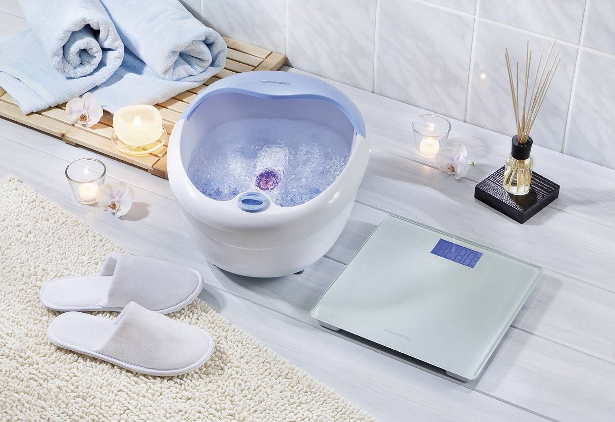 Bain de pieds avec un bord bleu, rempli d'eau et doté d'une fonction massage. Entouré de serviettes, de bougies et d'un pèse-personne sur un sol en bois clair.