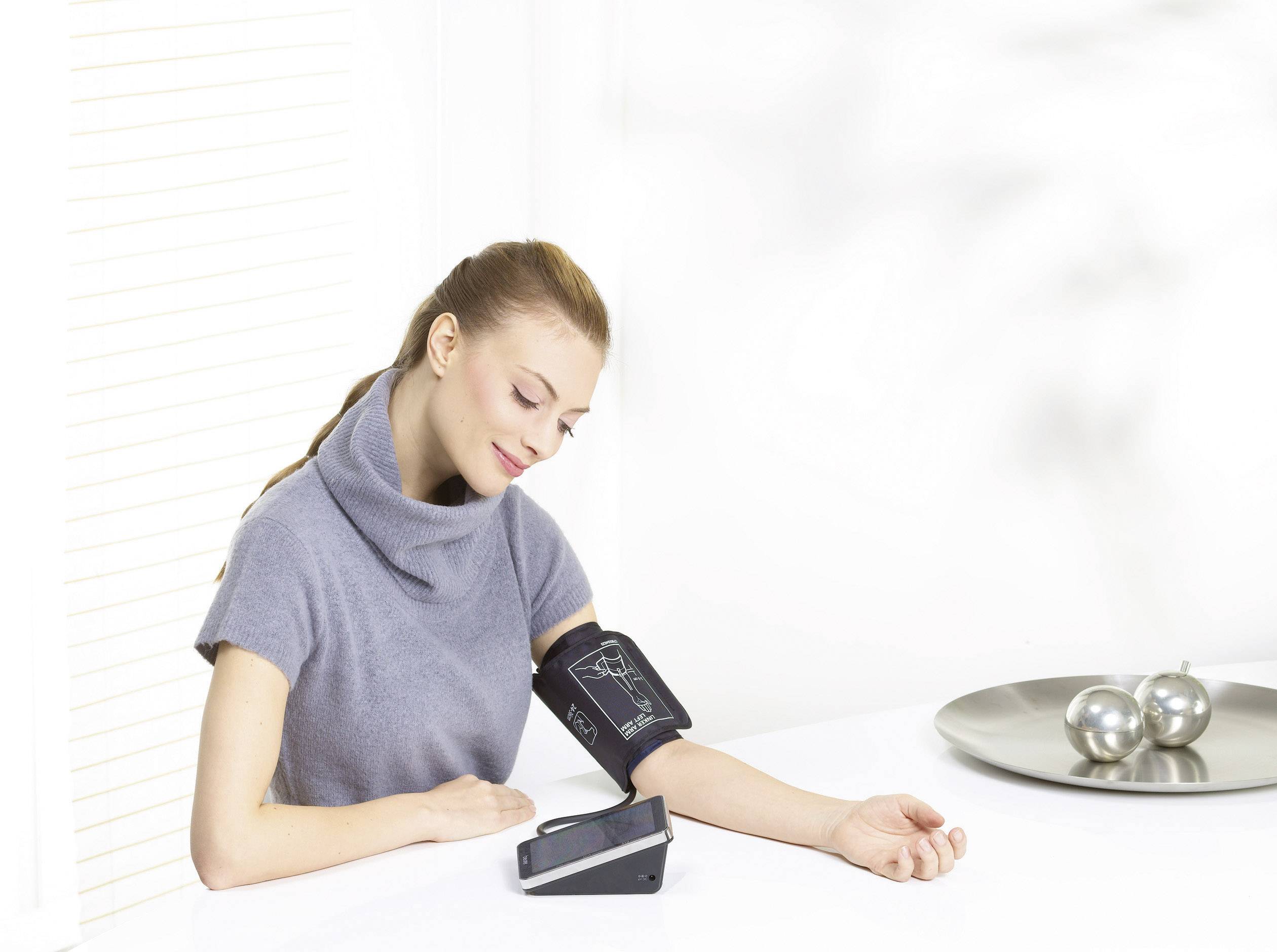 Une femme mesure sa tension artérielle avec un tensiomètre électronique alors qu'elle est assise à une table. Elle a l'air satisfaite.