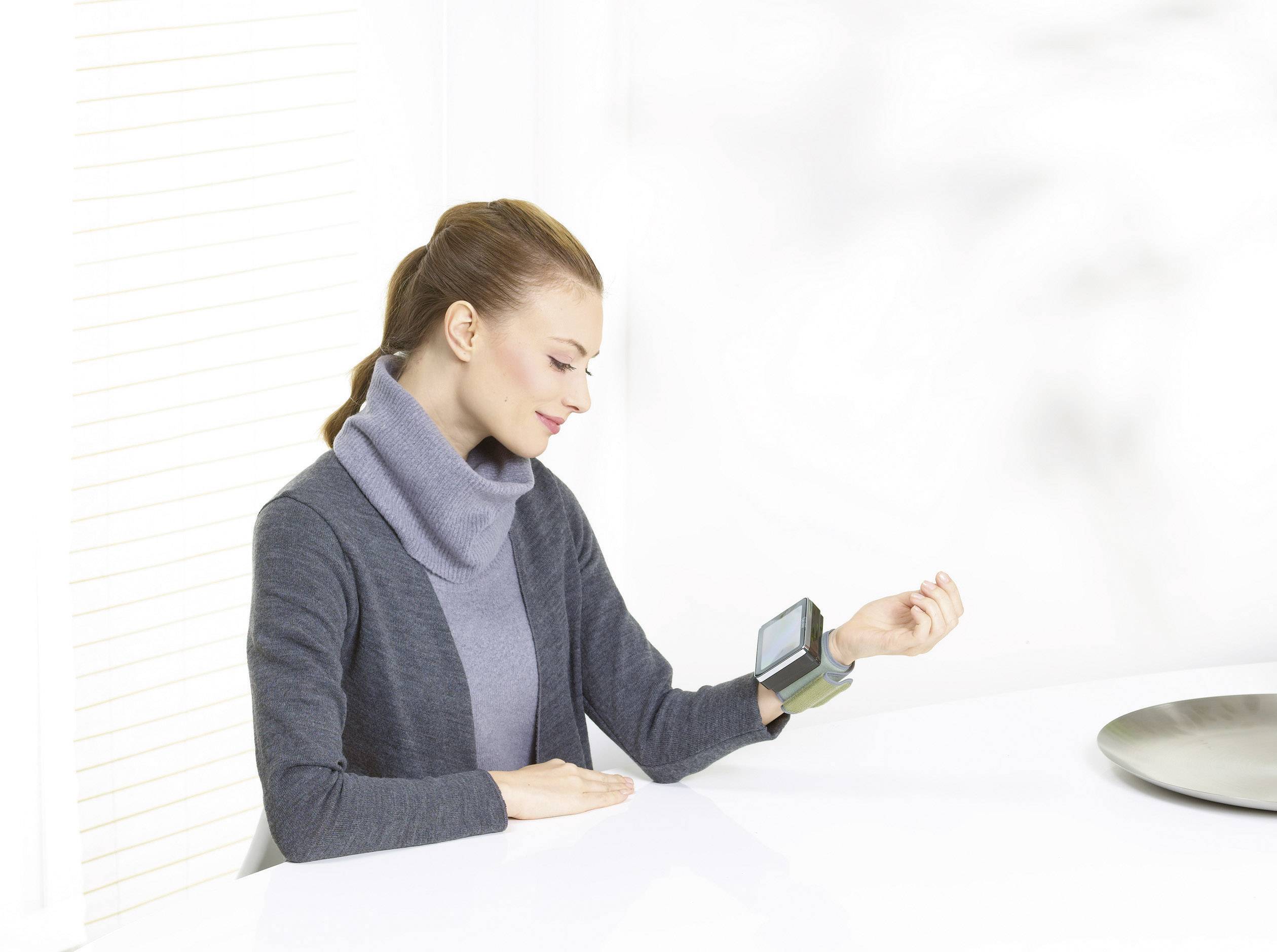 Une femme est assise à une table et mesure sa tension artérielle à l'aide d'un appareil numérique de mesure de la pression artérielle au poignet.