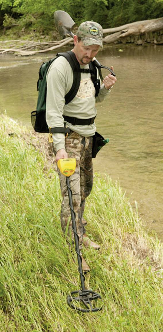 Un homme utilise un détecteur de métaux sur la berge d'une rivière, vêtu de façon décontractée et portant un camouflage, regardant attentivement le sol.