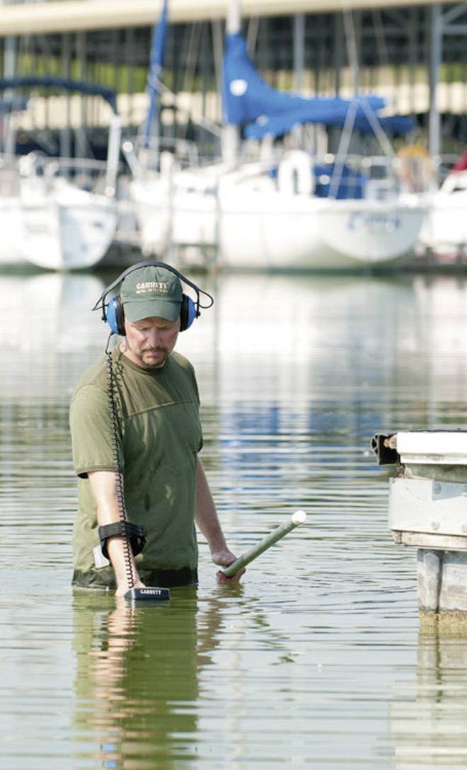 Un homme marche dans l'eau avec un détecteur de métaux près d'un port. Des bateaux sont visibles en arrière-plan.