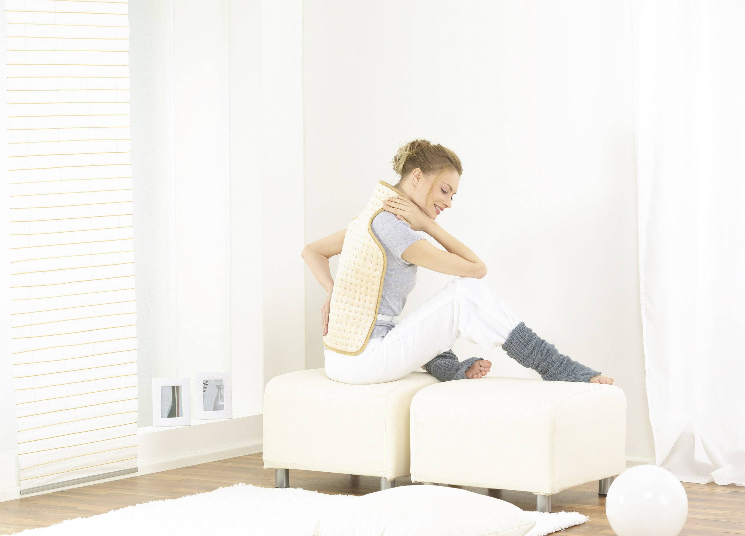 Une femme est assise sur un tabouret dans une pièce lumineuse, portant un gilet chauffant. Elle a l'air détendue et tient sa main sur sa nuque.