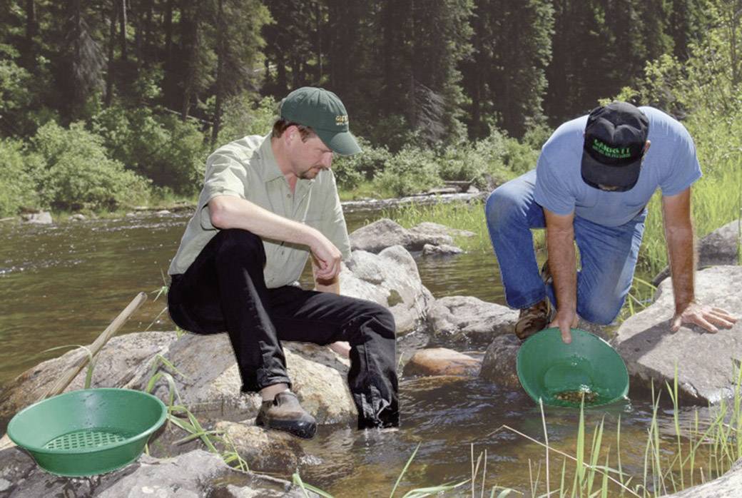 Deux hommes sont assis au bord d'une rivière et cherchent de l'or dans des plateaux verts. Les environs sont boisés.