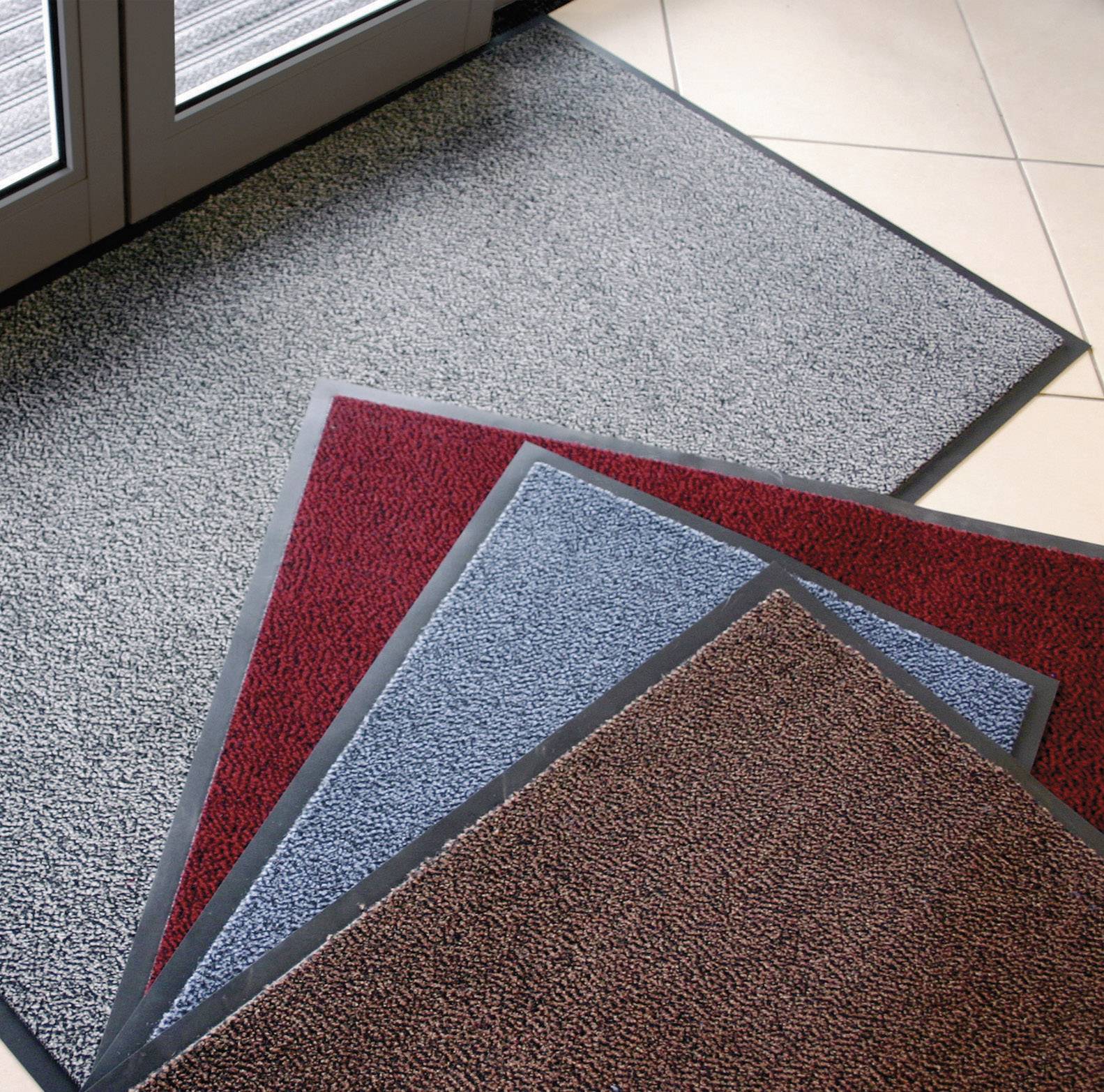 Différents tapis de sol en gris, rouge, bleu et marron sont superposés sur un carrelage devant une porte vitrée.