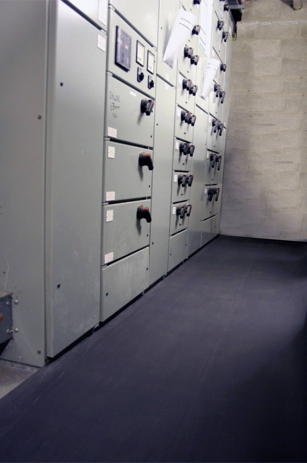 Rangée de panneaux de commande gris dans une salle technique. Plusieurs interrupteurs à bascule sont visibles. Le sol est recouvert d'un tapis noir.