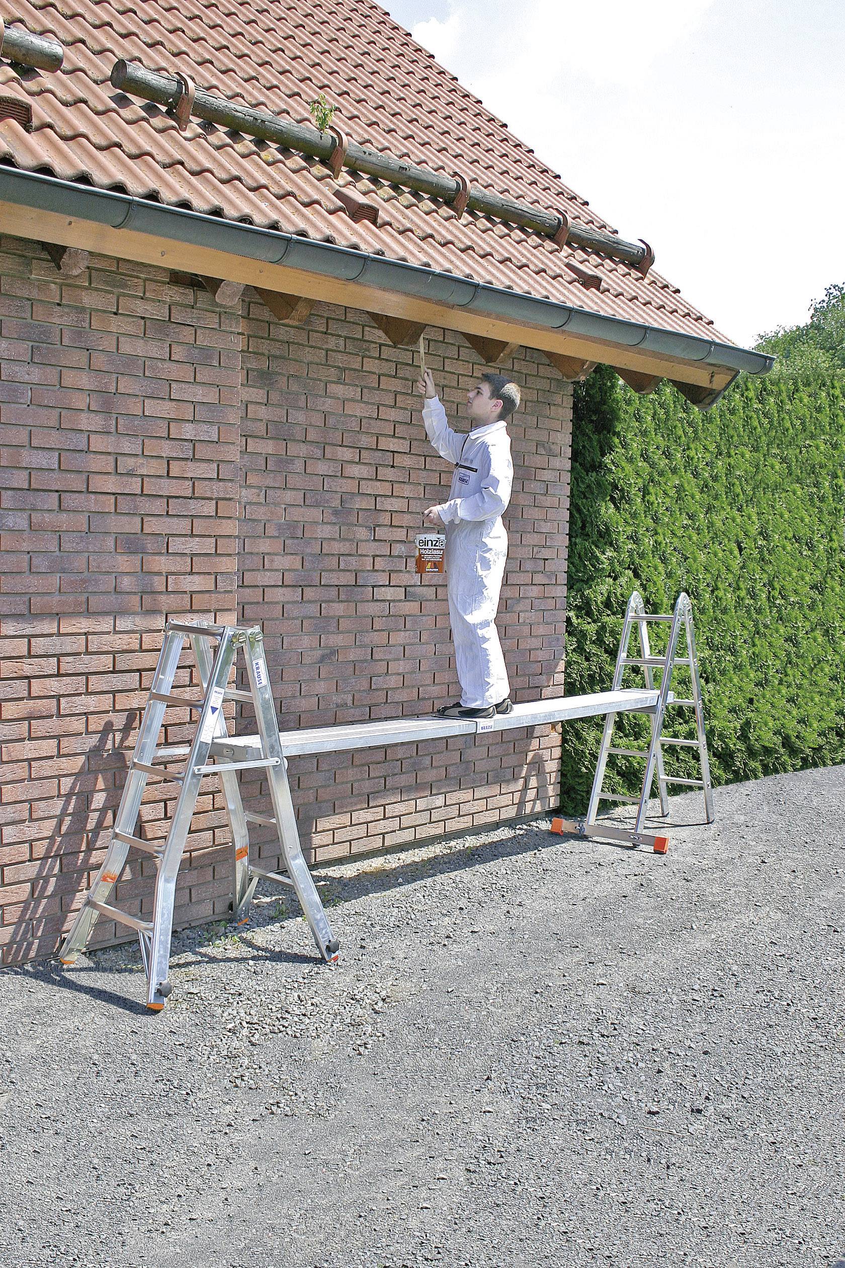 Une personne en combinaison blanche est debout sur un échafaudage de fortune composé de deux échelles et d'une planche et peint la gouttière d'une maison.