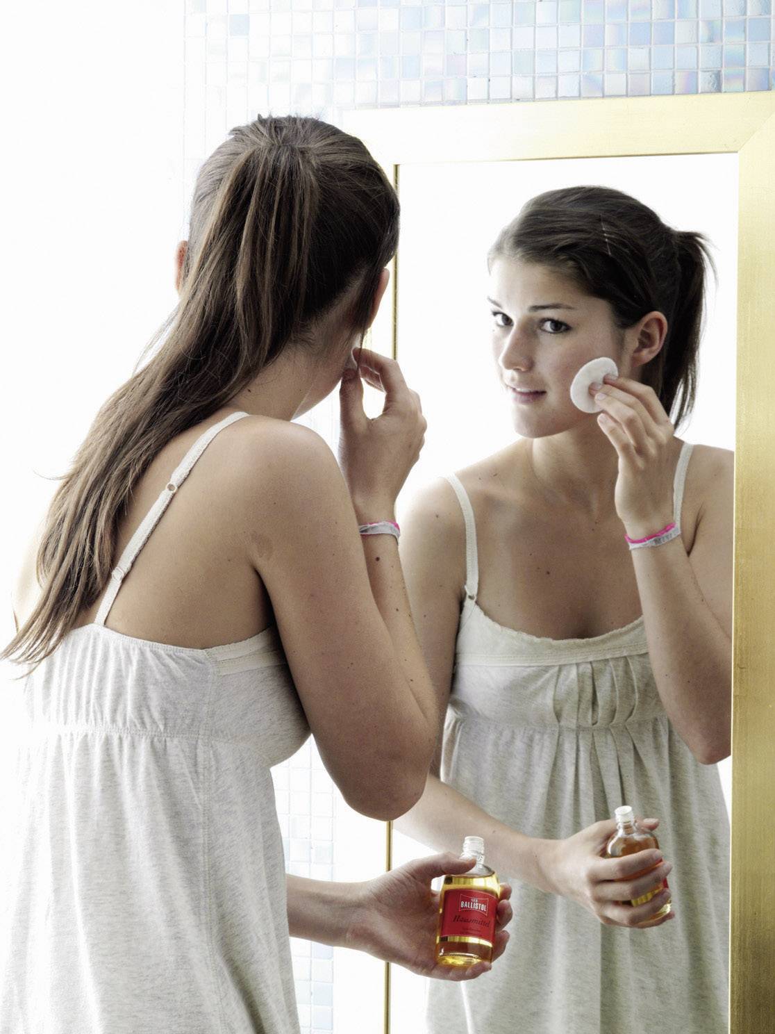 Une femme se tient devant un miroir et nettoie son visage avec un coton démaquillant, une bouteille de démaquillant à la main.