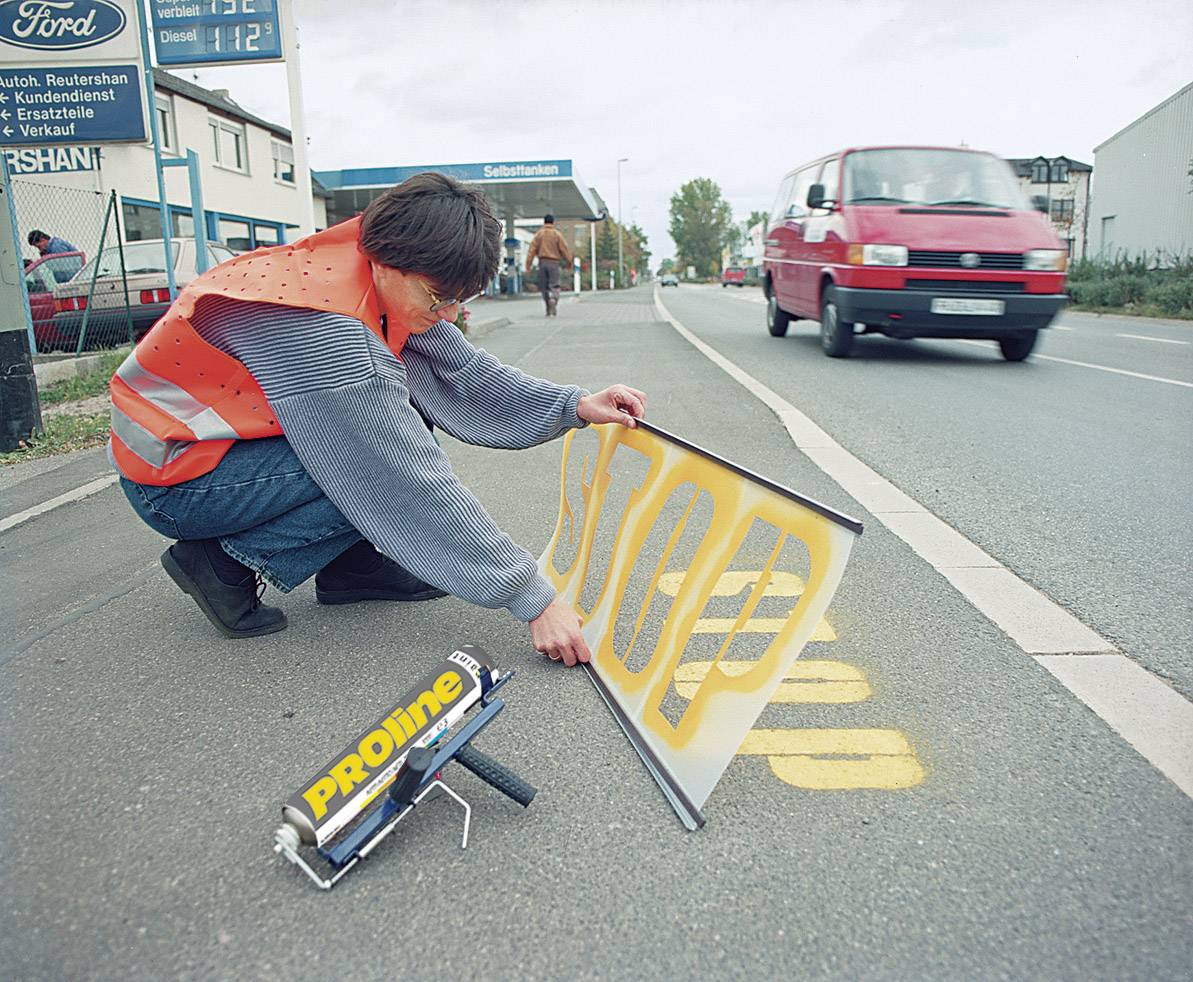 Un ouvrier en vêtements de sécurité écrit le mot 'STOP' sur la route à l'aide d'un pochoir, alors qu'une voiture rouge passe à proximité.
