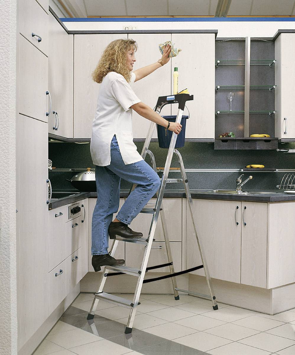 Femme sur une échelle en train de nettoyer les placards de cuisine supérieurs ; portant des vêtements décontractés ; cuisine moderne avec du bois clair et des plans de travail sombres.