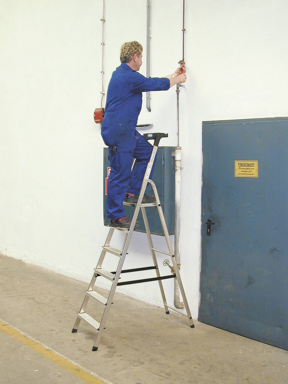 Une personne vêtue de bleu, portant des vêtements de travail, est debout sur une échelle et effectue des réparations sur une conduite de tuyauterie contre le mur.