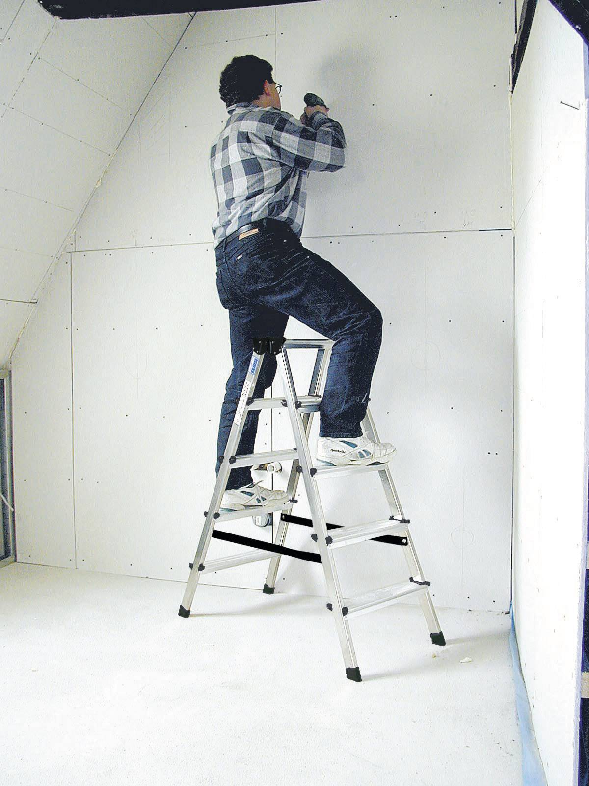 'Une personne fixe des plaques de plâtre sur un mur, debout sur une échelle dans une pièce inachevée, portant une chemise à carreaux et un jean.'