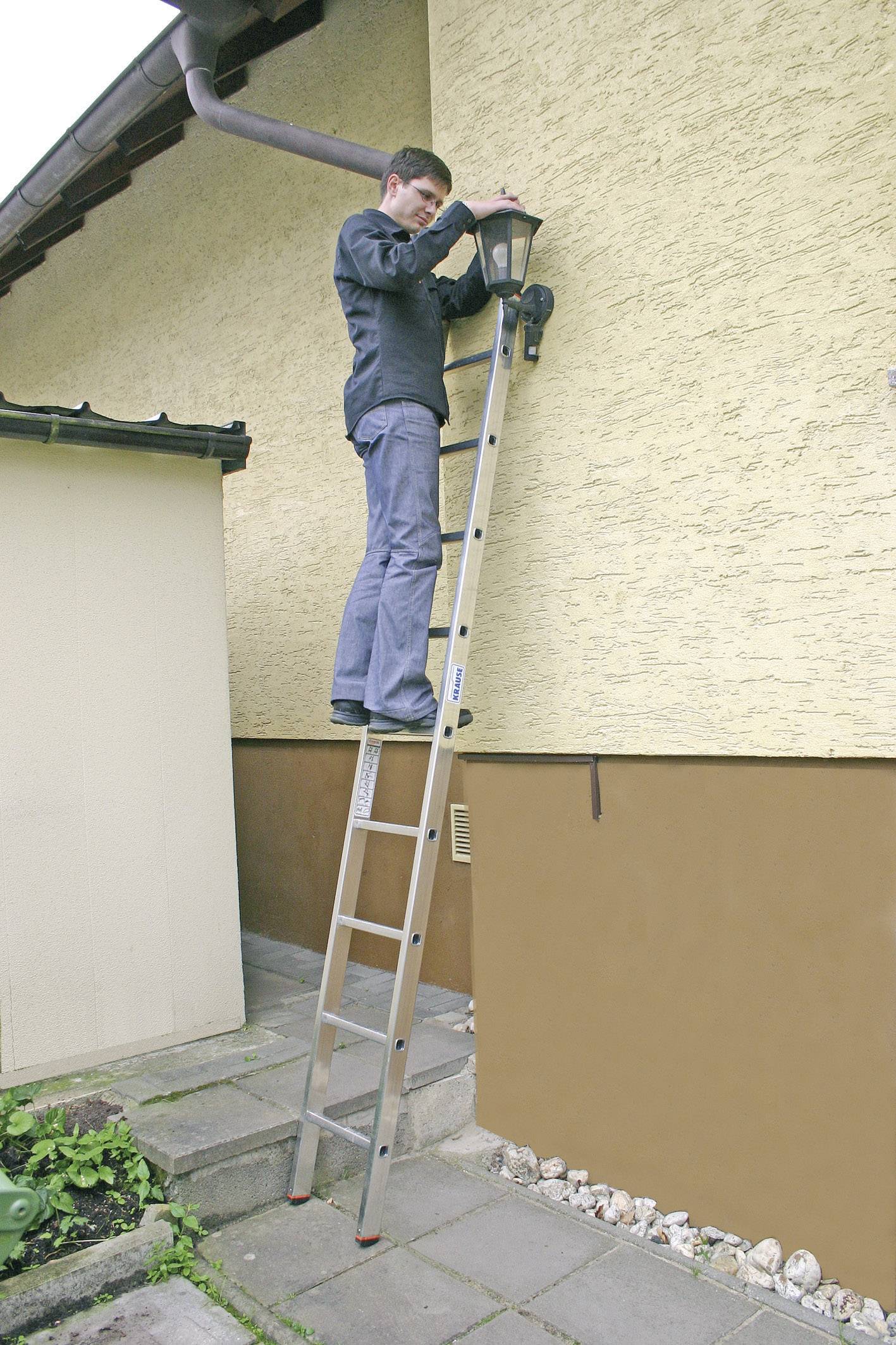 Un homme est debout sur une échelle et répare une lampe extérieure sur le mur d'une maison.