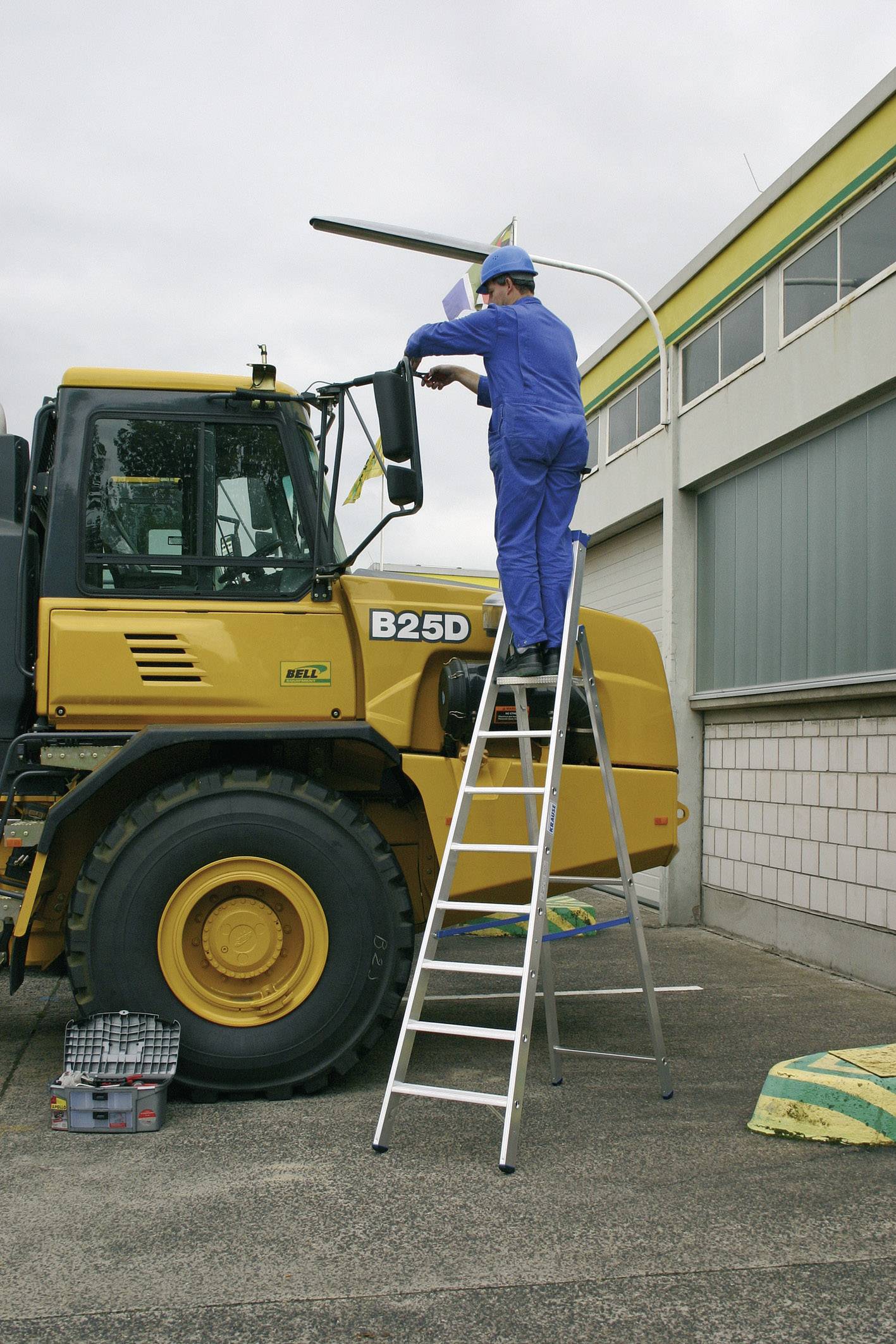 Un ouvrier en tenue de travail bleue est debout sur une échelle et répare un lampadaire à côté d'un véhicule de chantier jaune.