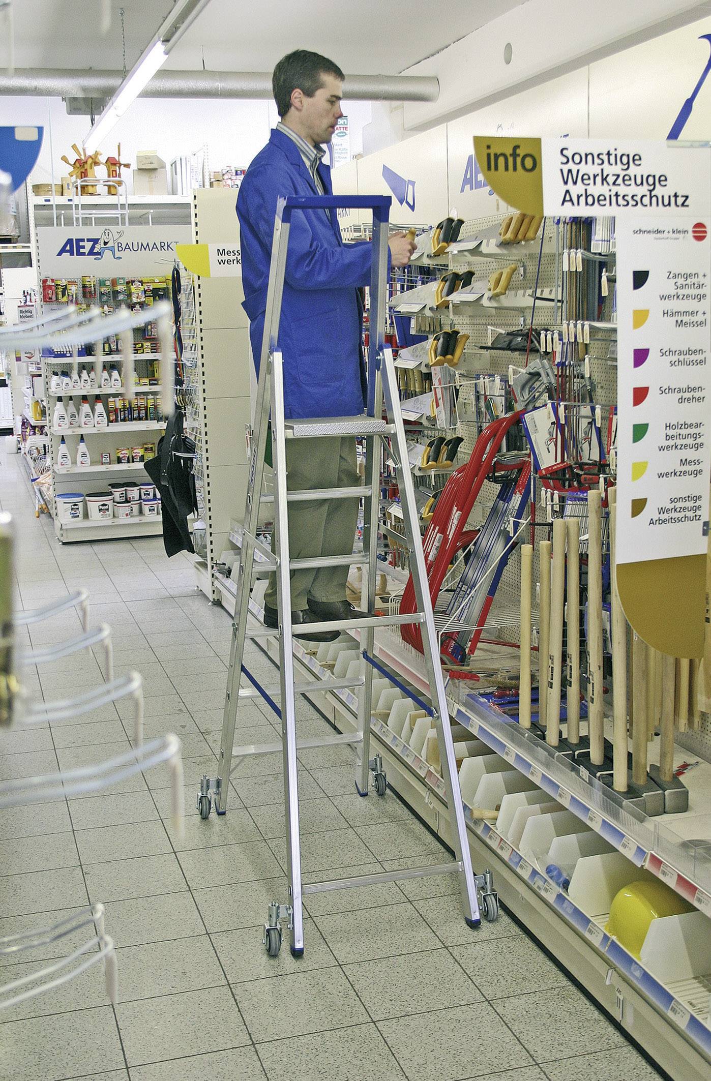 Une personne se tient sur un escabeau dans un magasin de bricolage et range des outils. Des étagères avec divers articles sont visibles en arrière-plan à gauche.